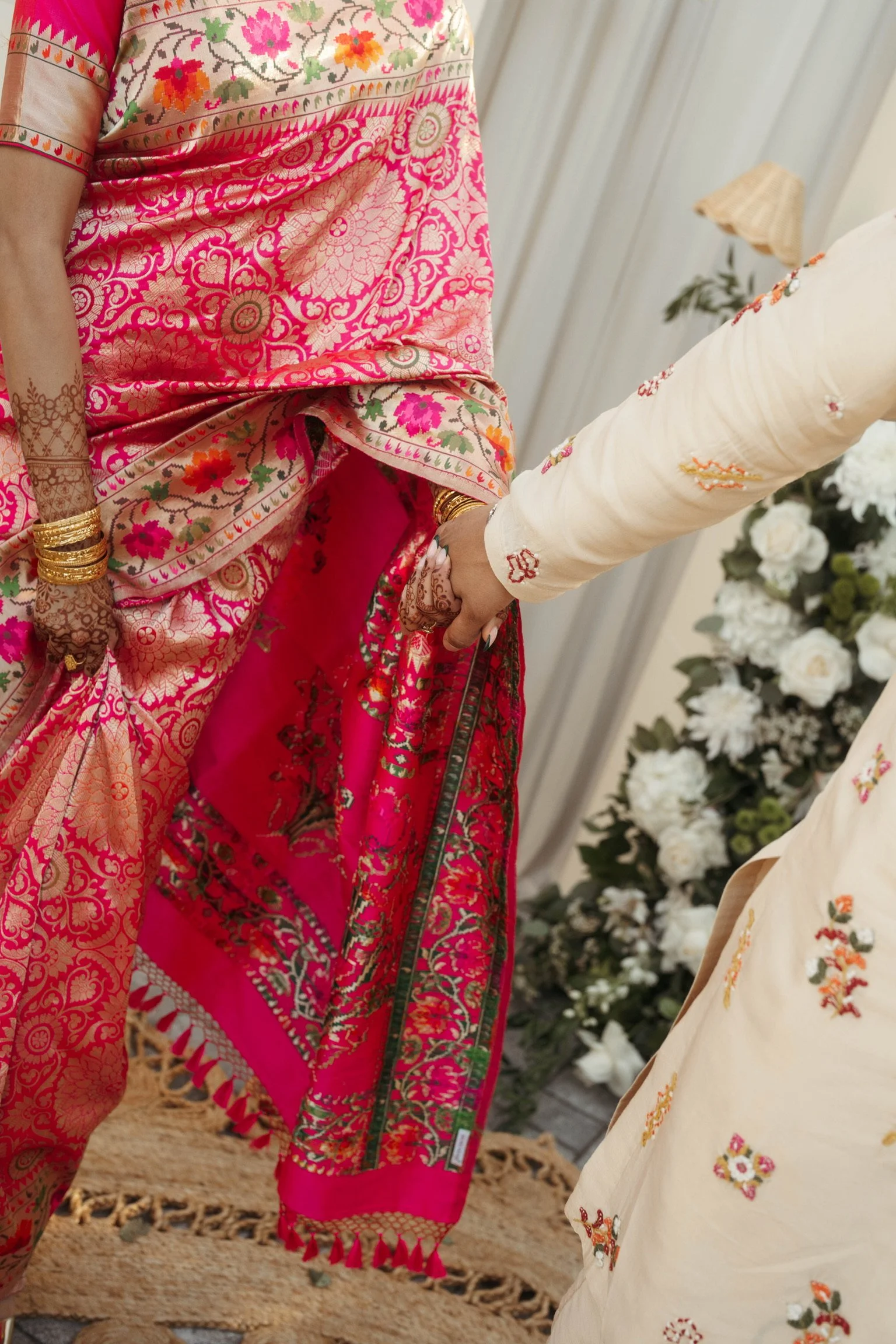 Close-up of two women wearing colorful traditional Indian saris, holding hands, with flowers and a woven rug in the background.