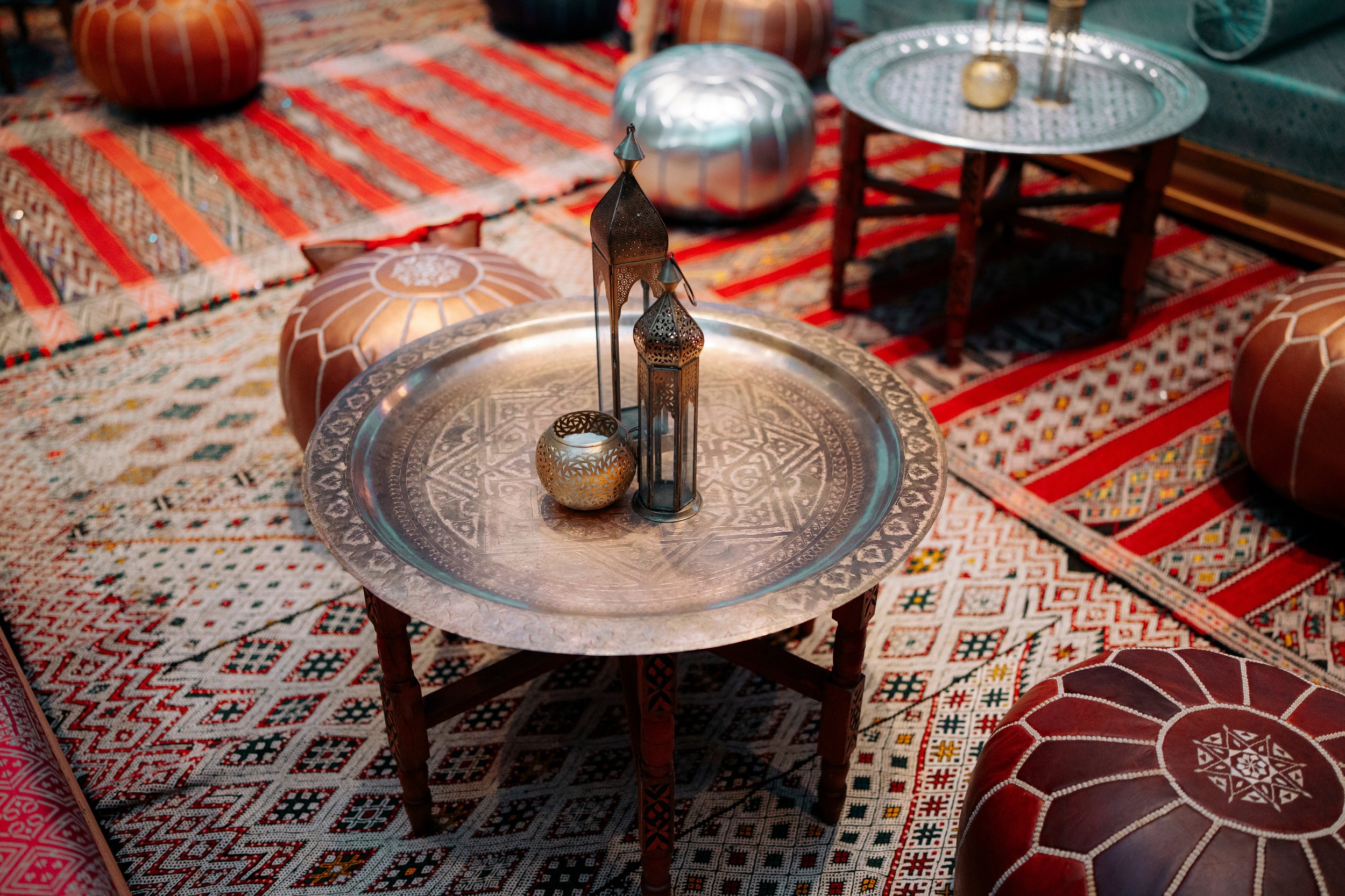 Decorative Middle Eastern-style room with a silver tray table, candles, lanterns, and patterned cushions on a vibrant, intricate area rug.