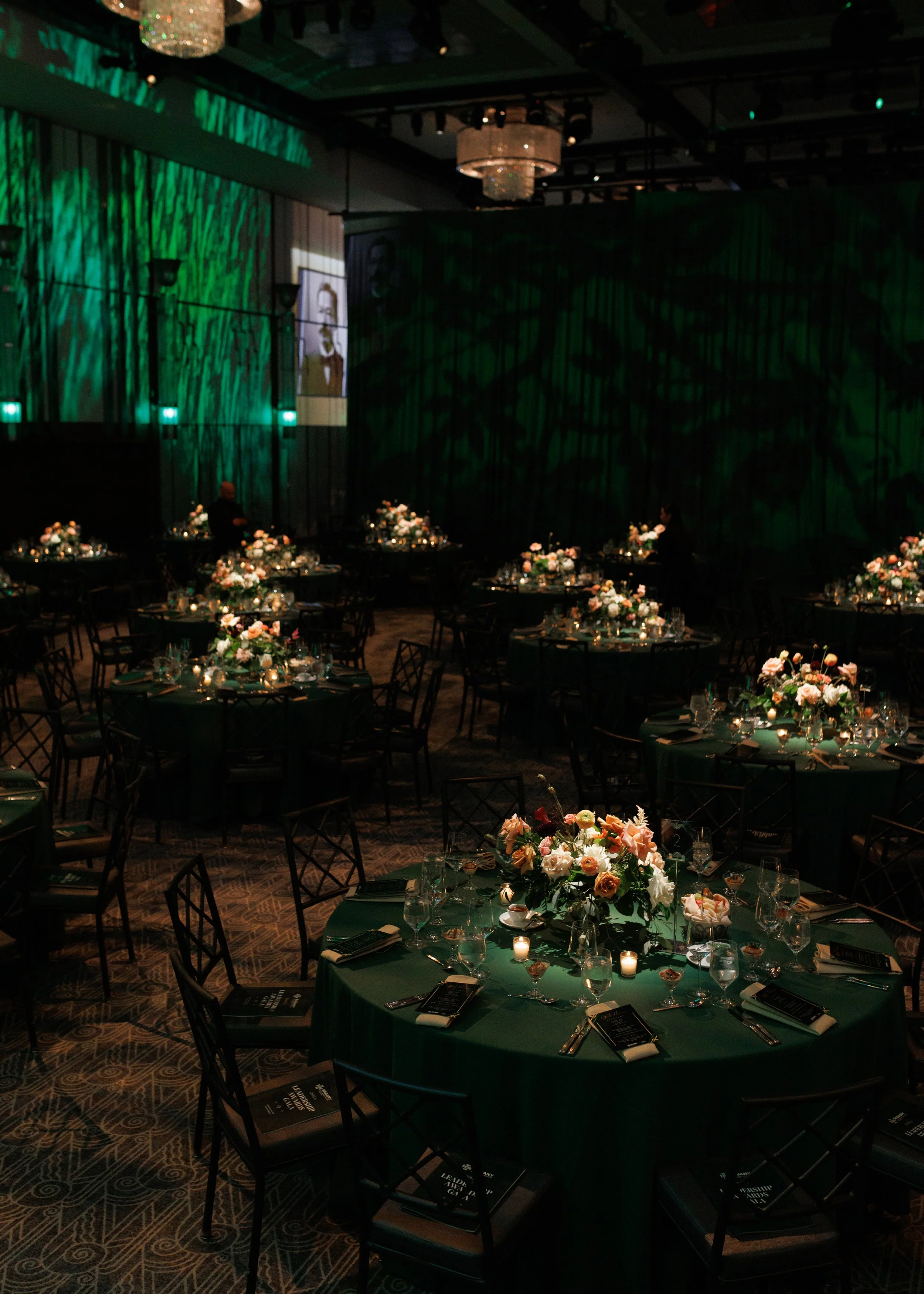 Elegant banquet hall with round tables covered in dark green tablecloths, each decorated with floral centerpieces, candles, and place settings, dimly lit with green uplighting and chandeliers.