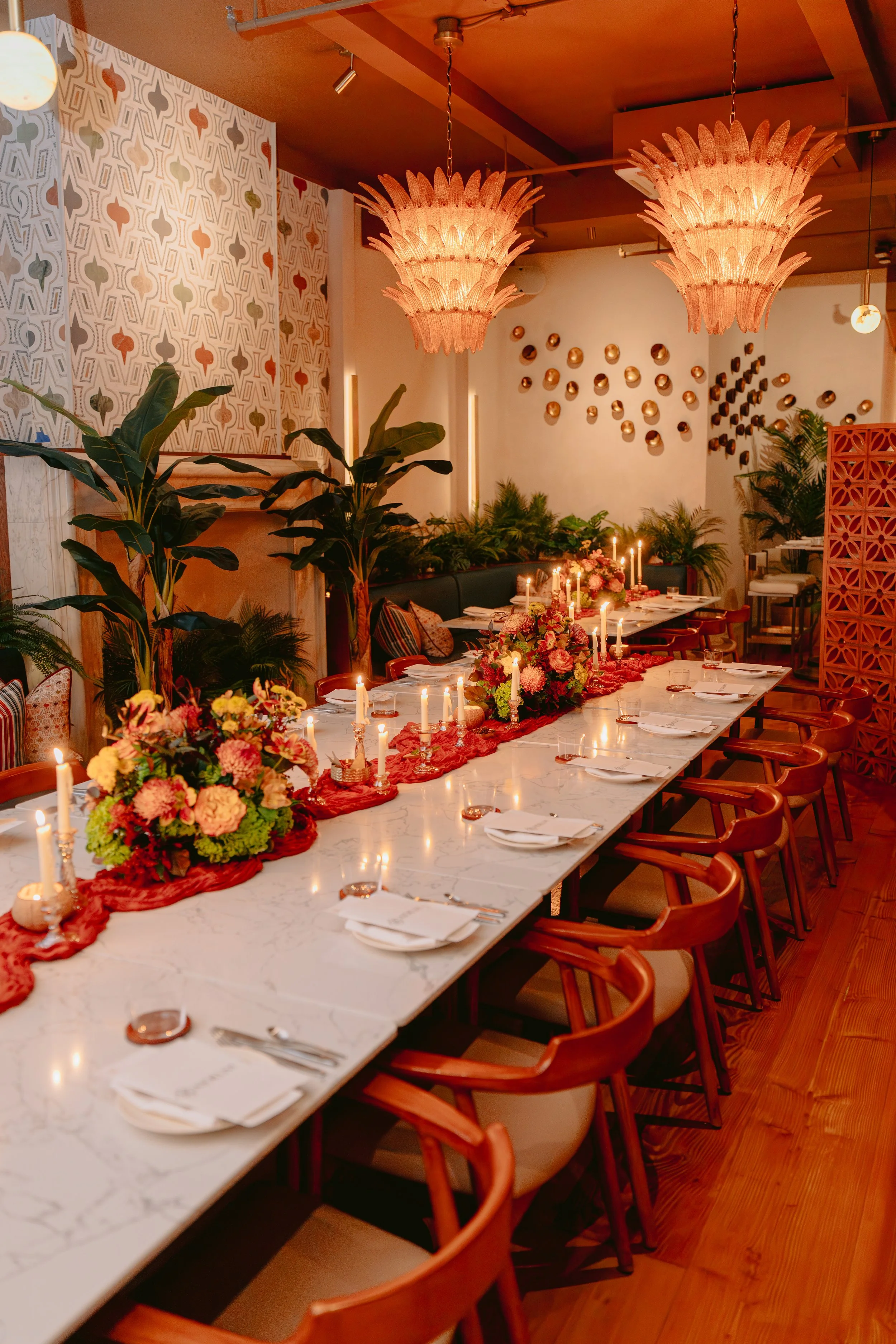Decorated dinner table with floral arrangements, candles, and place settings inside a warmly lit restaurant with wall art and hanging lamps.