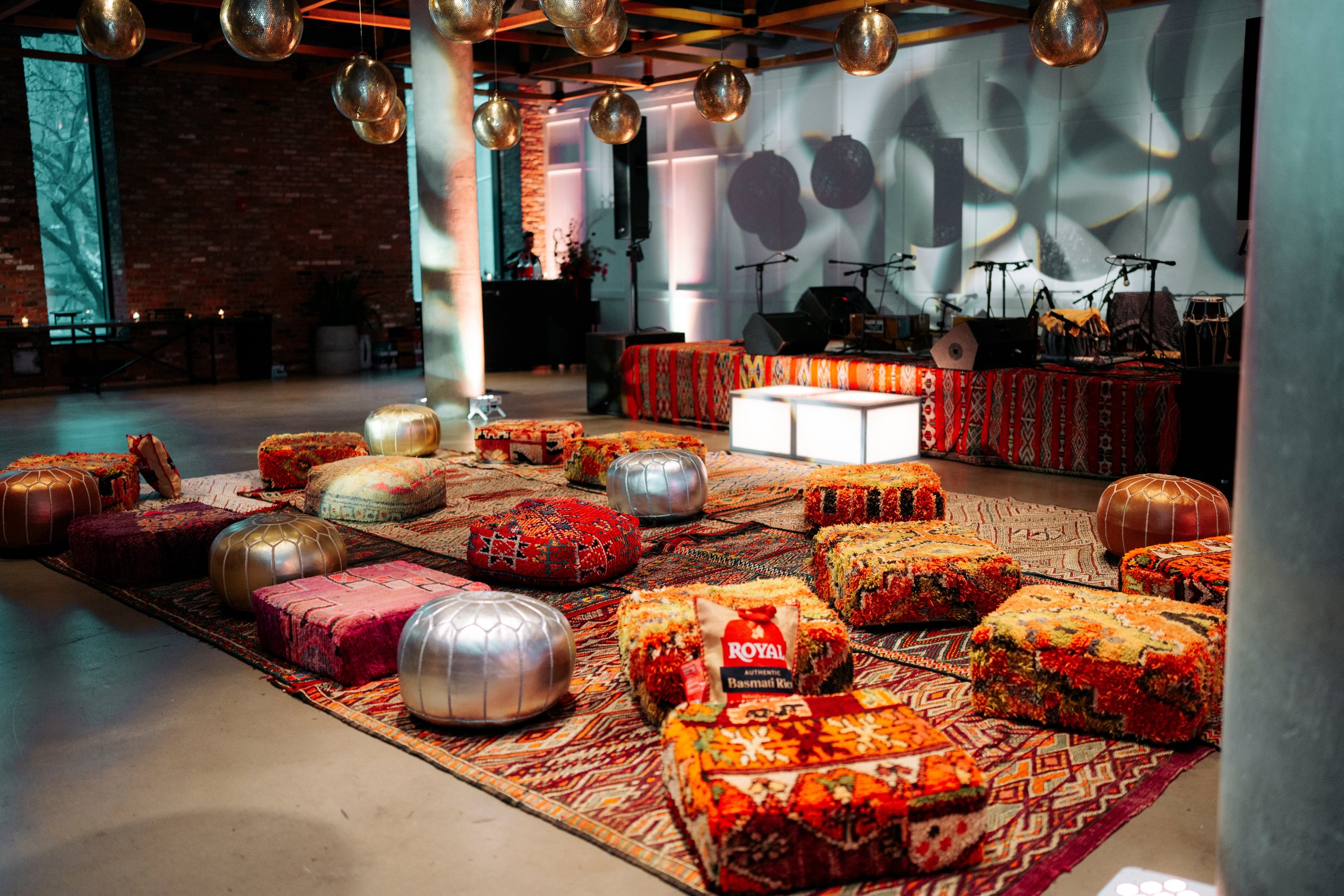 A cozy event space decorated with colorful Persian rugs, woven cushions, and metallic poufs in front of a stage with microphones and musical instruments, illuminated with soft lighting, featuring a brick wall and large windows.