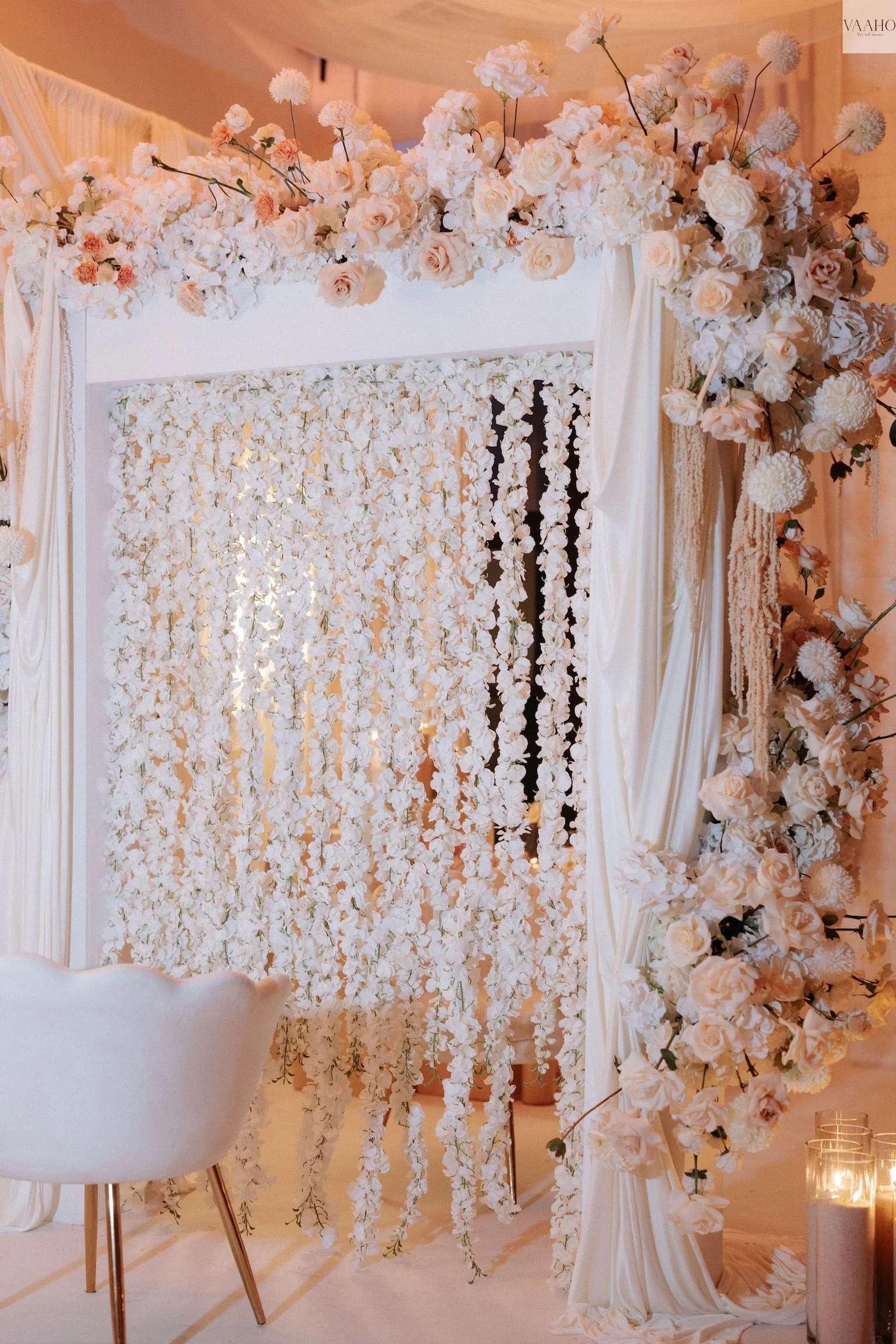 A decorative floral backdrop with white and blush roses, hydrangeas, and other flowers, arranged around a white draped fabric and a mirror with hanging floral strands, in warm lighting.