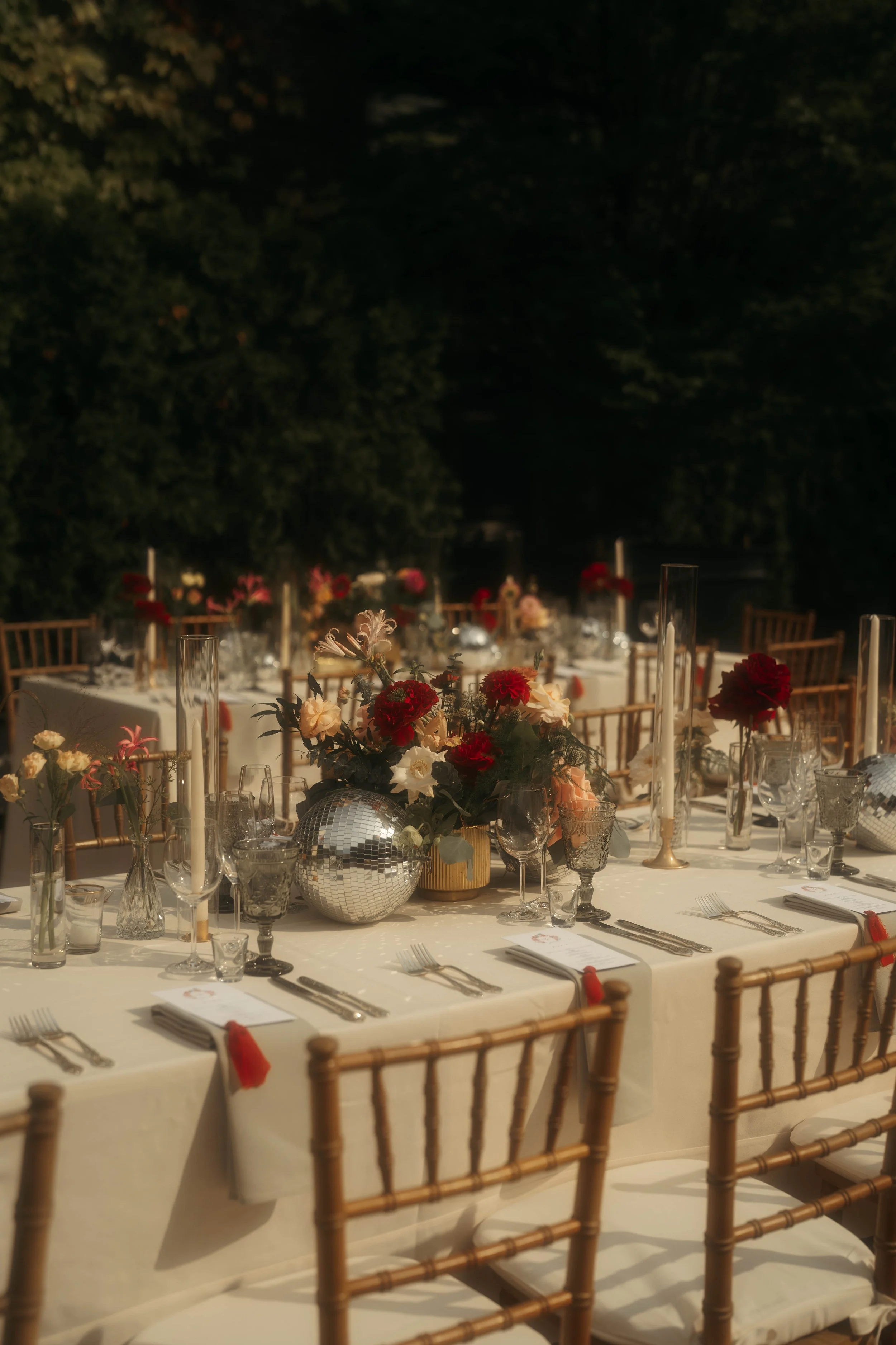 An outdoor banquet table decorated with floral centerpieces, glassware, candles, and table settings, set for an evening event with chairs around the table, under a tree at nighttime.