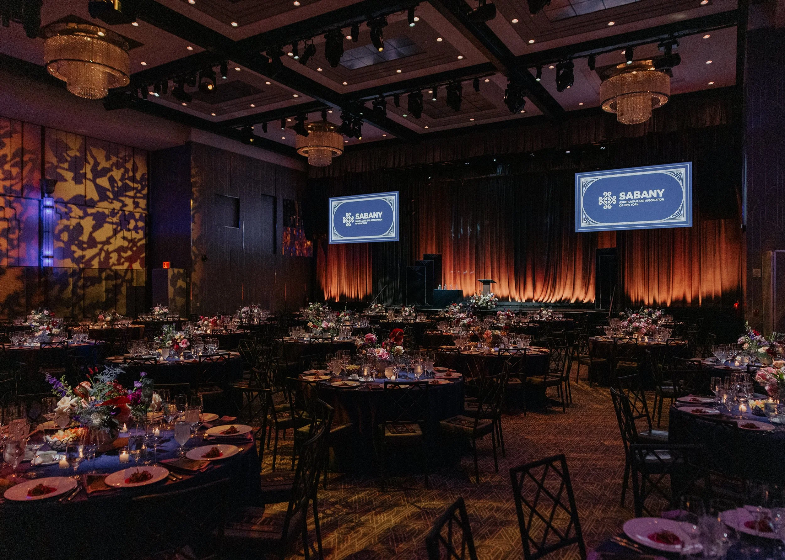 A large banquet hall with tables set for a formal event. The tables are decorated with flower arrangements and dinnerware, and the stage at the front displays a sign for the South Asian Bar Association of New York. The hall features elegant lighting,