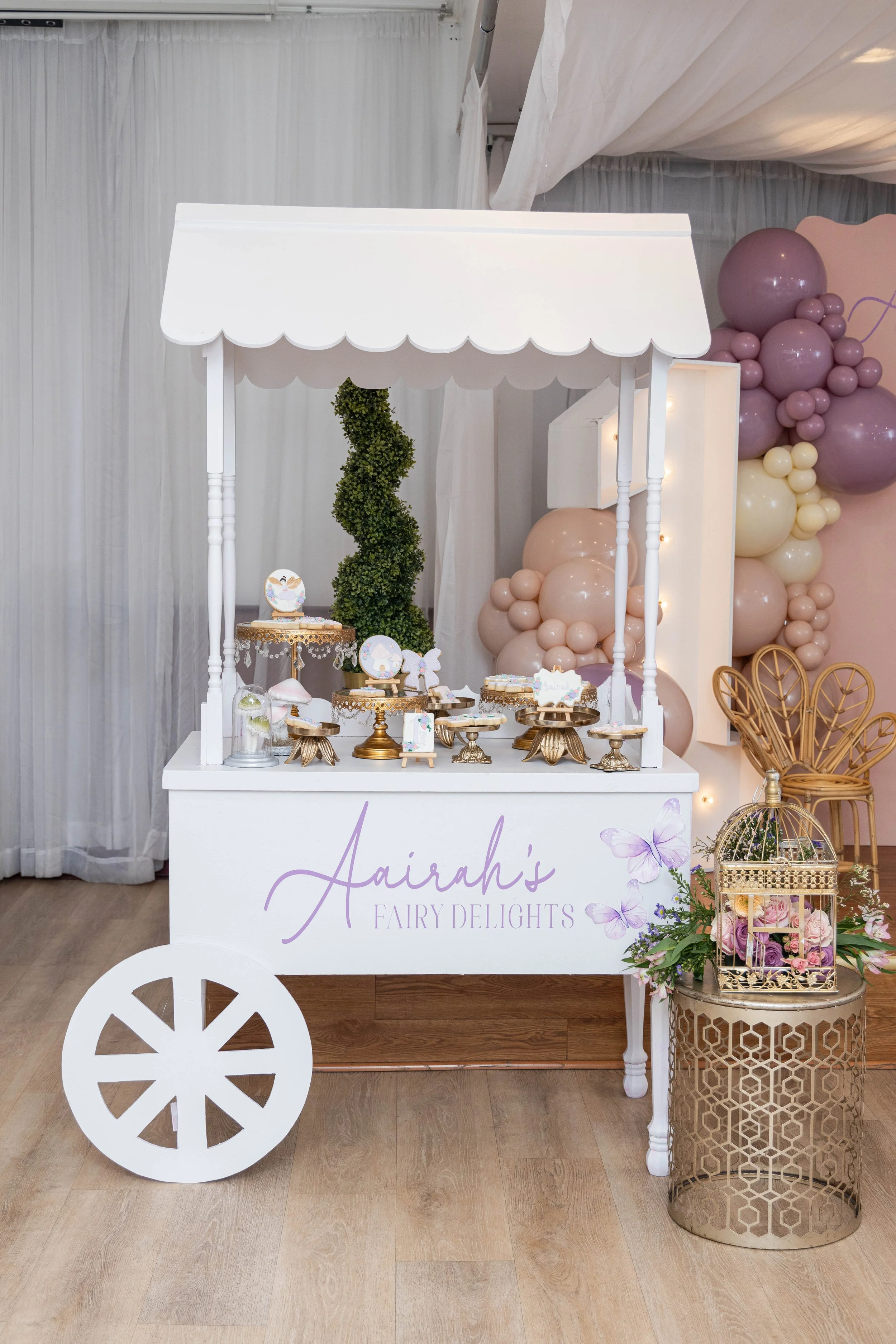 A dessert cart decorated for a celebration with a sign reading 'Aairah's Fairy Delights'. The cart is white with purple lettering and is filled with cupcakes and treats. To the right of the cart, there is a golden decorative table with pink and purpl
