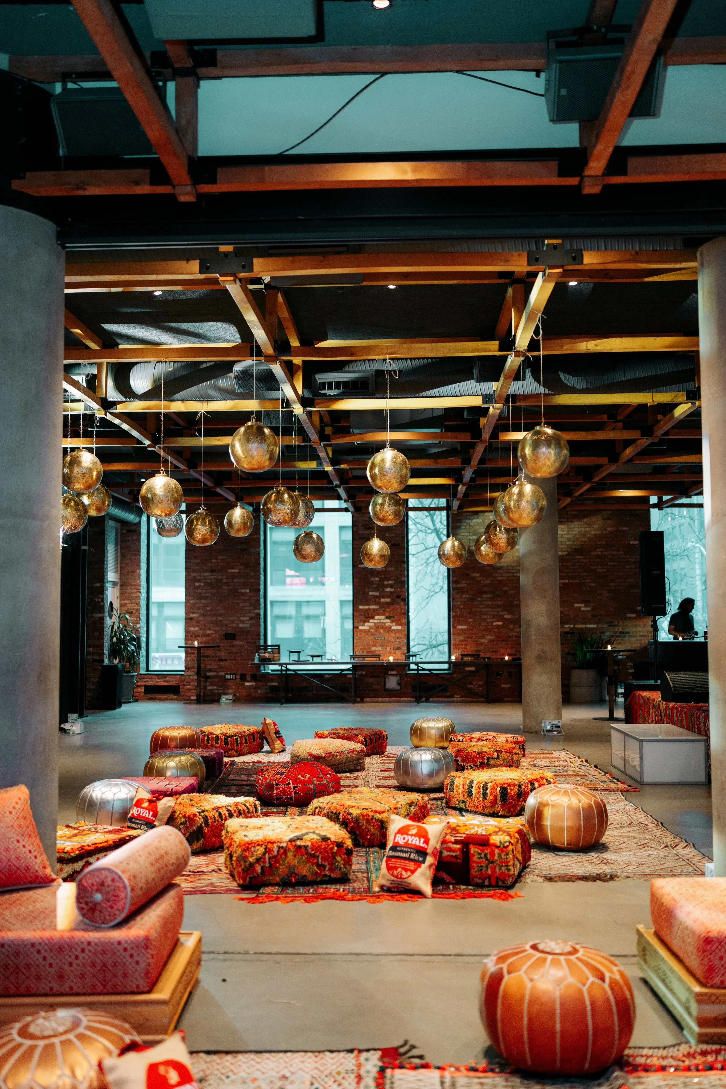 Interior of a room with a high ceiling and large windows. The ceiling has wooden grid structures, and metallic lanterns shaped like globes hang from it. The floor is decorated with various patterned carpets, cushions, and poufs, creating a cozy loung