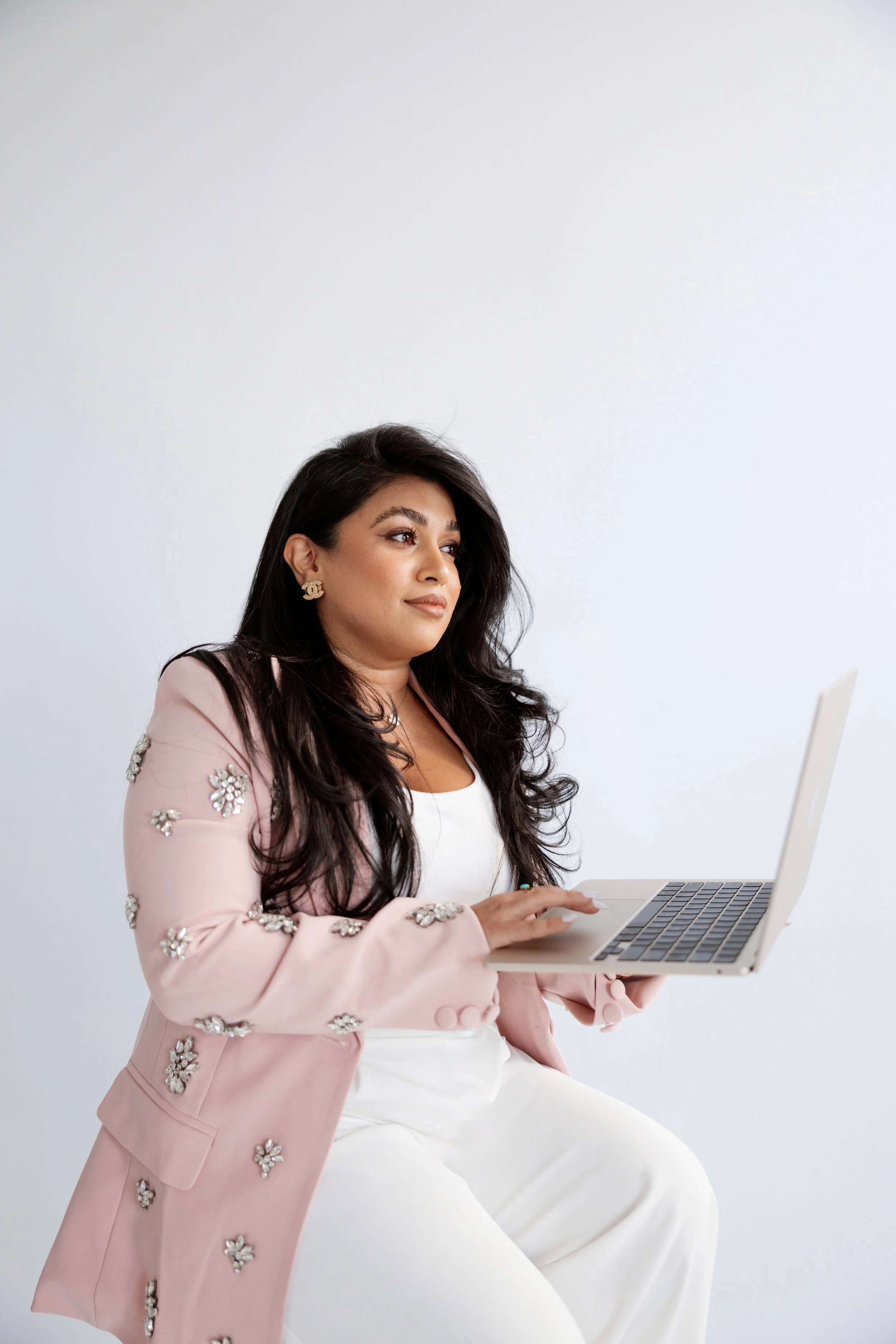 A woman sitting, holding a laptop, wearing a pink blazer with floral embellishments, and white pants, against a plain white background.