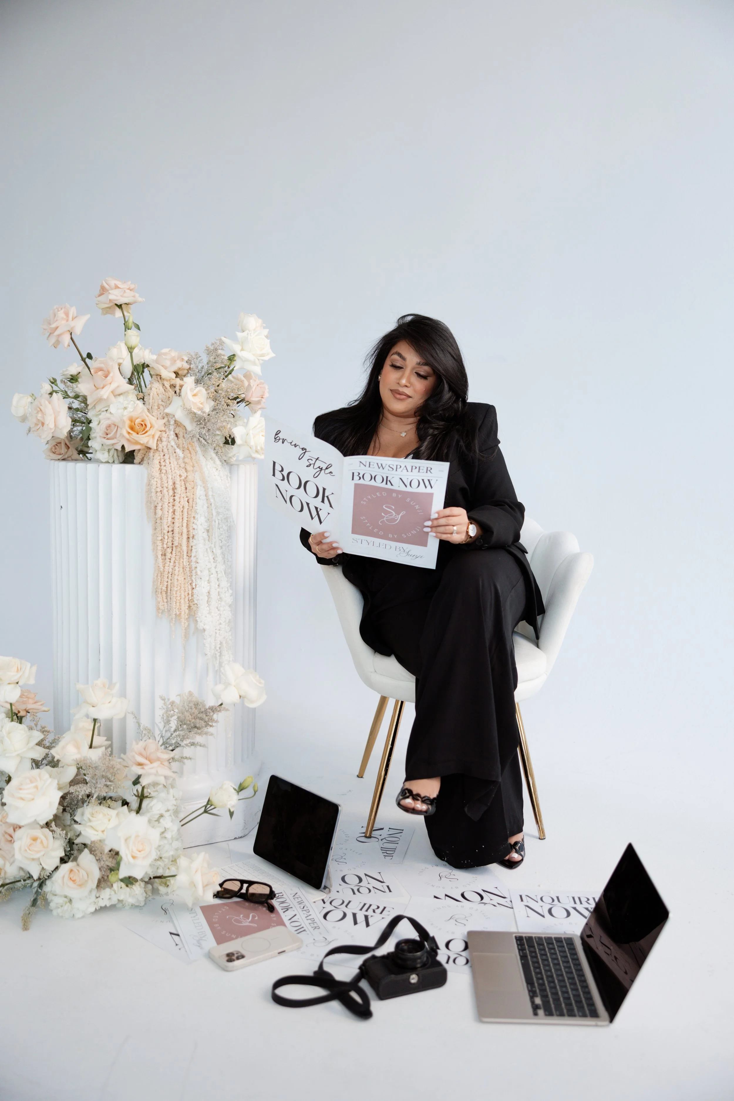 A woman sitting on a white chair, reading a magazine, surrounded by flowers, electronics, and printed papers on the floor, with a plain white wall in the background.