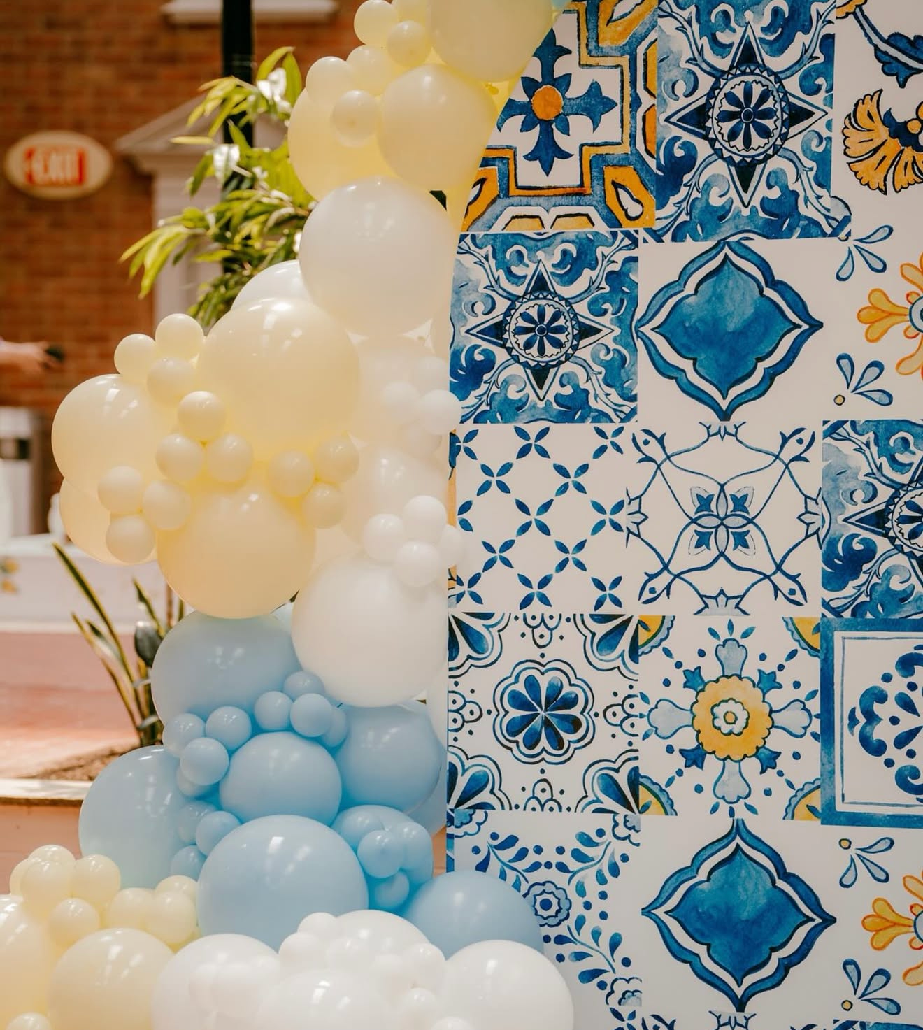 Decorative wall with blue and yellow tile patterns and a balloon arrangement in pastel colors including white, cream, and light blue, with some greenery in the background.