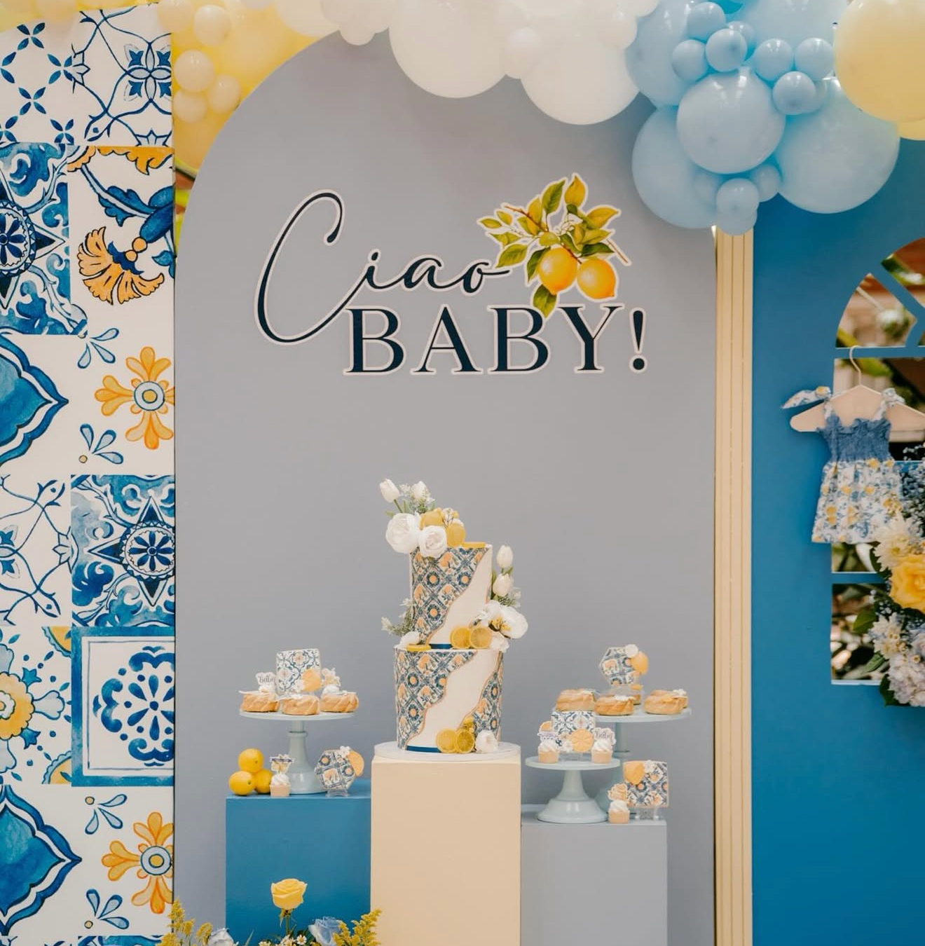 Decorative setup for a baby celebration with a light blue background, a cake, cupcakes, lemons, and a sign that says "Ciao Baby!" in black lettering, surrounded by blue and white balloons and floral decorations.