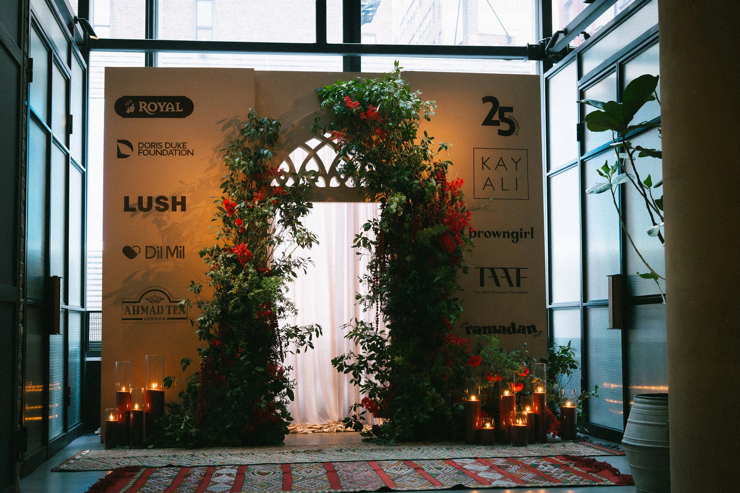 Indoor event space with floral and candle decorations, patterned rug, and a backdrop with various sponsor logos including Royal, Doris Duke Foundation, Lush, Dil Mil, and others.