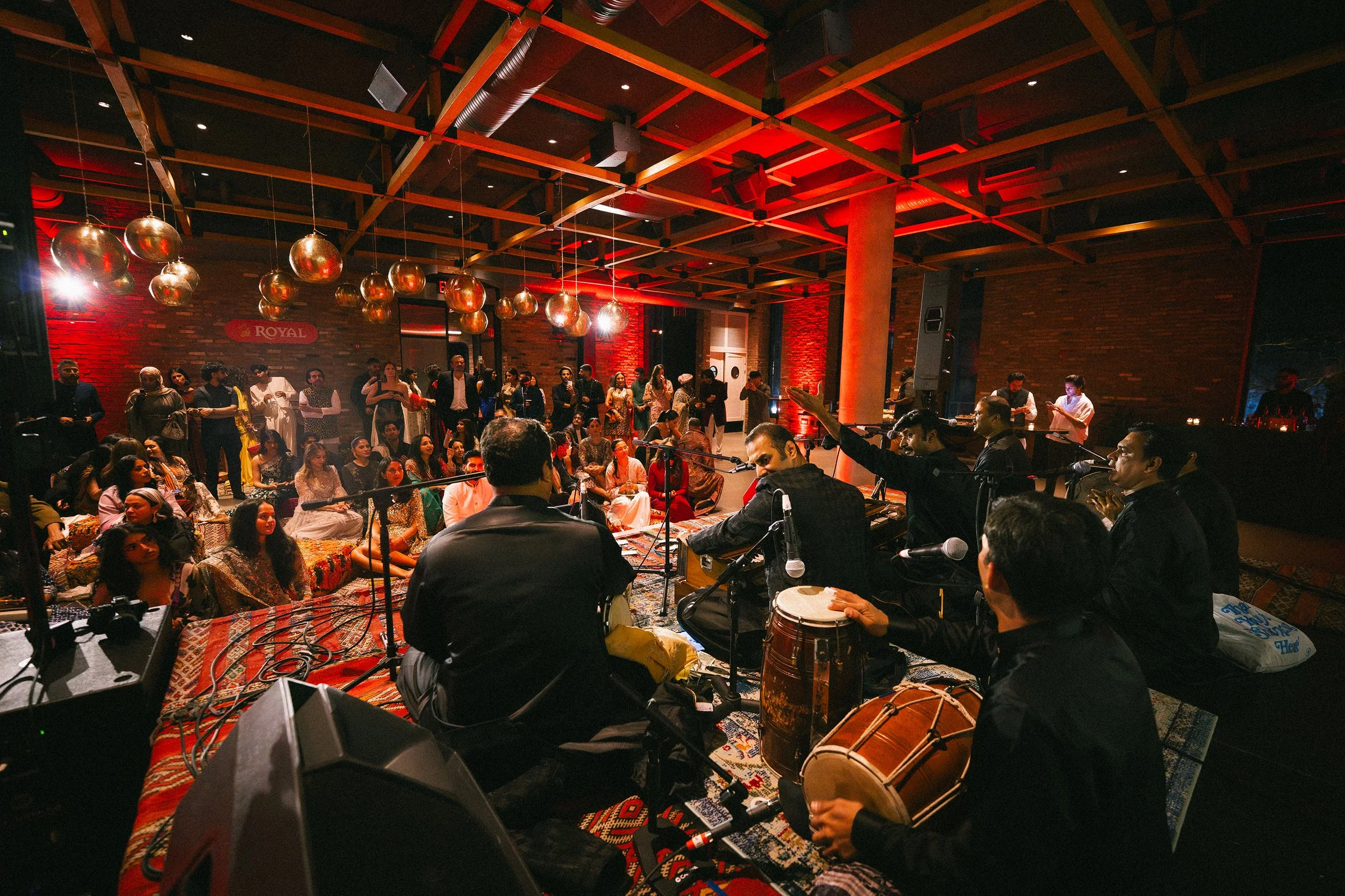An indoor event or concert with people sitting and standing, a stage with musicians playing traditional Indian instruments, and festive decor including red lighting and hanging gold spheres.