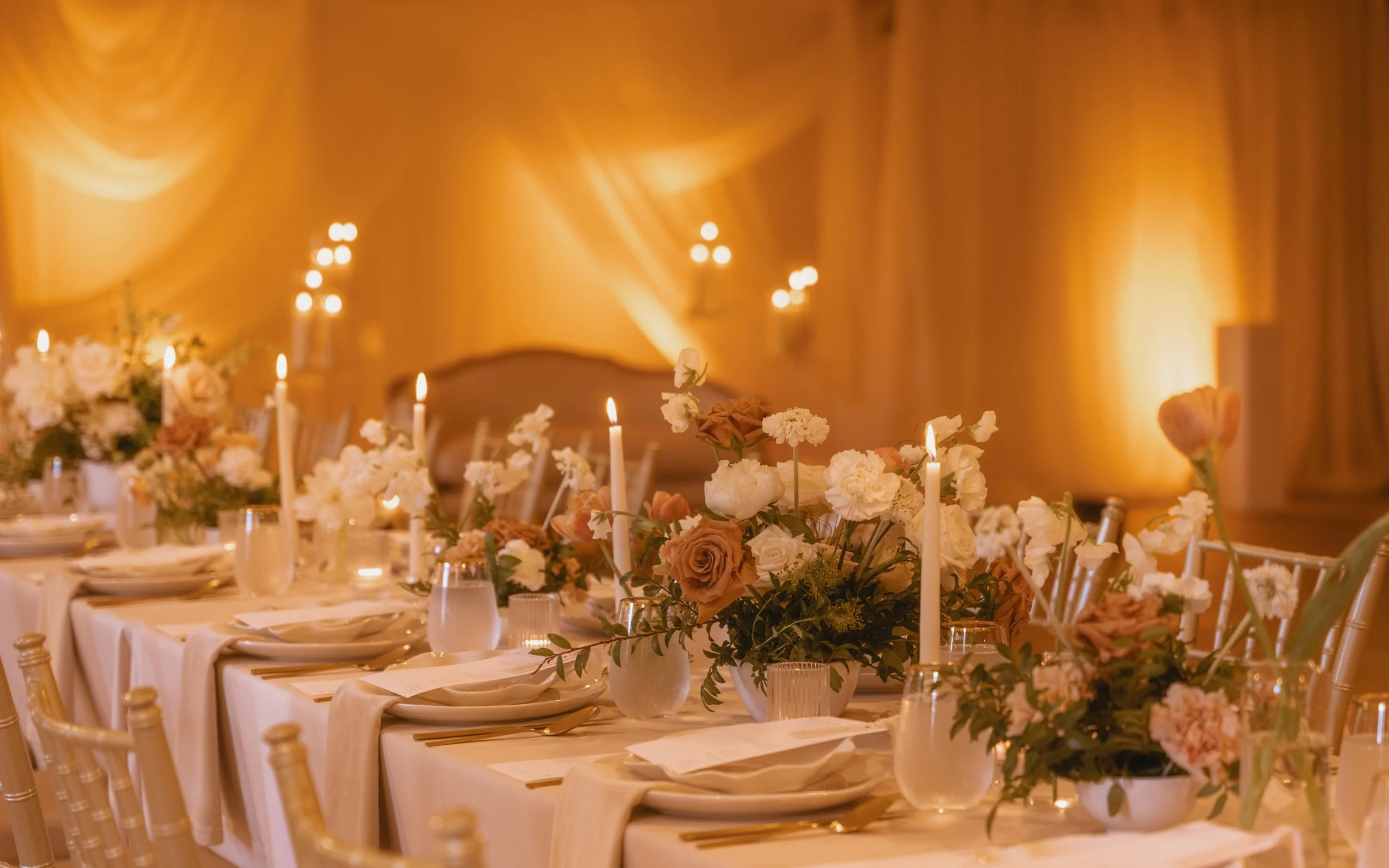 Elegant banquet table with floral centerpieces, white tablecloths, and candlelit decor in a warmly lit room.