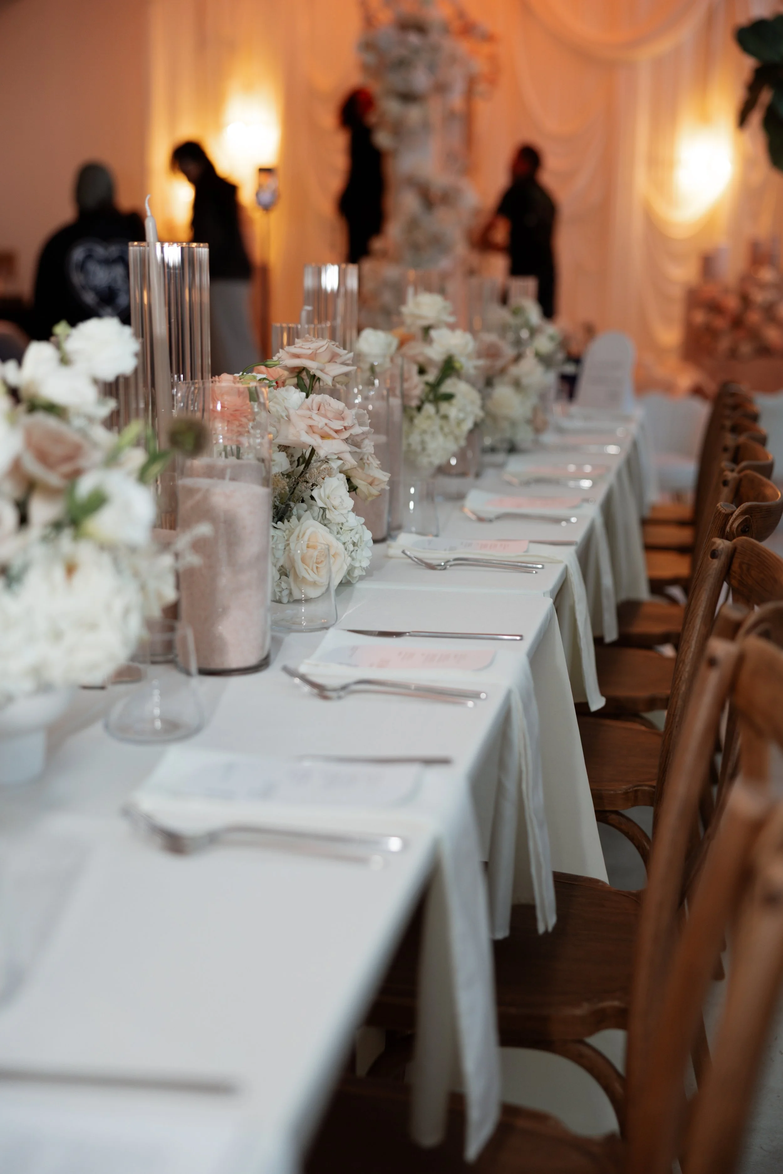 A long banquet table decorated with white and blush pink flowers in glass vases, with candles and place settings, set for an elegant event in a warmly lit room. People are in the background, standing and chatting.