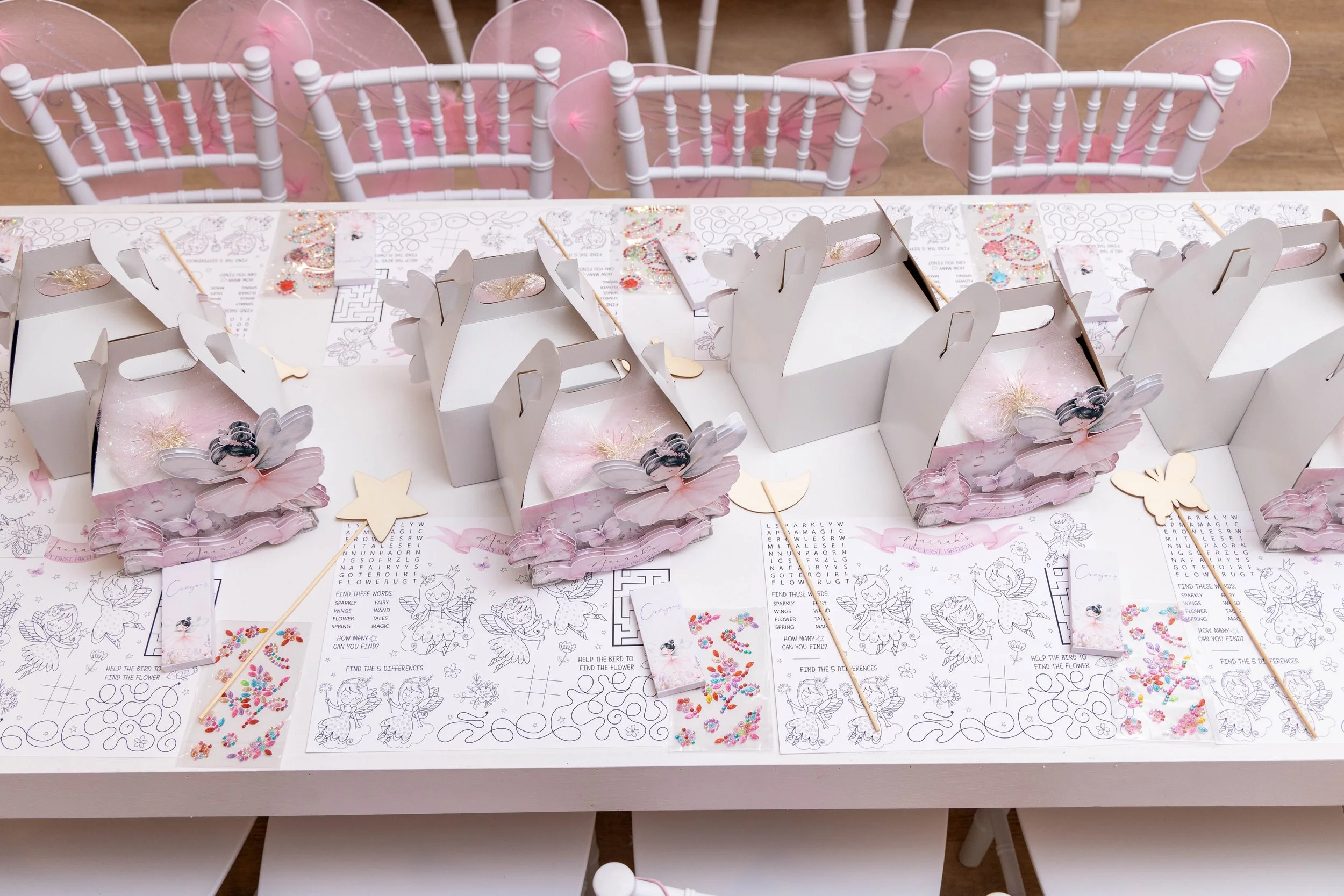 Table decorated with pink fairy-themed party favors and activity sheets, surrounded by white chairs with pink fairy wings decorations.