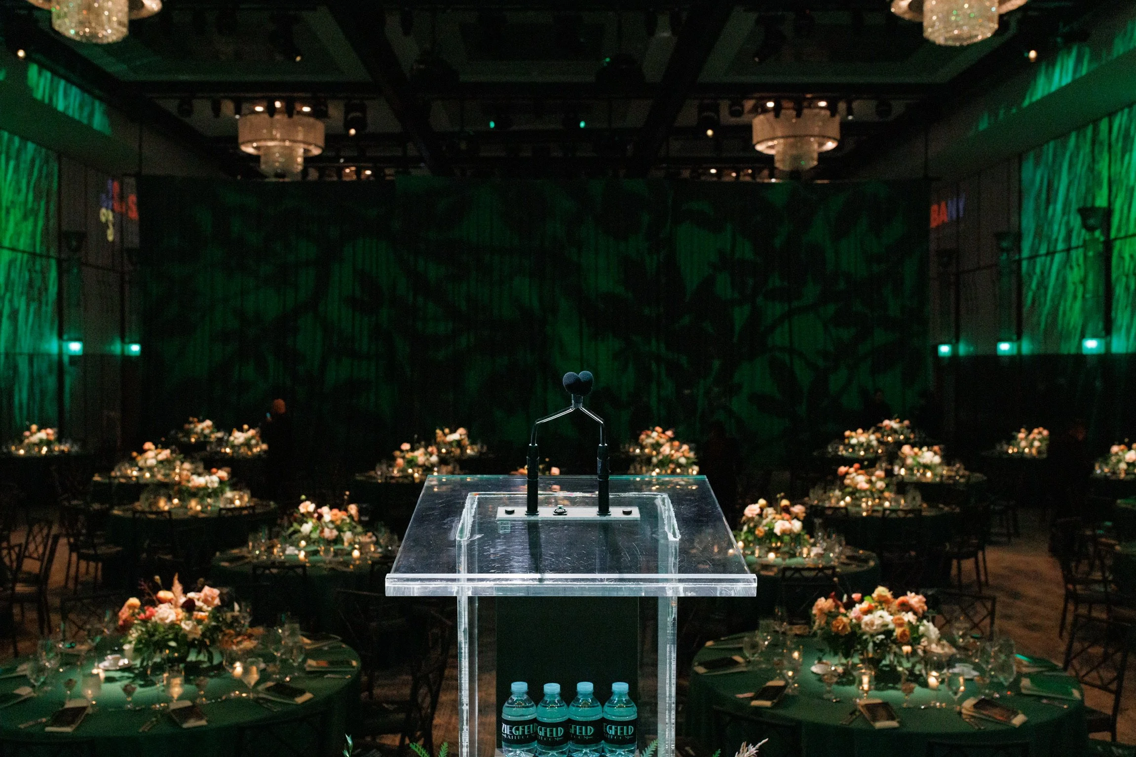 A clear podium with a microphone in the foreground at a decorated event venue, with tables set with flowers, candles, and bottled water, and a large stage with green lighting in the background.