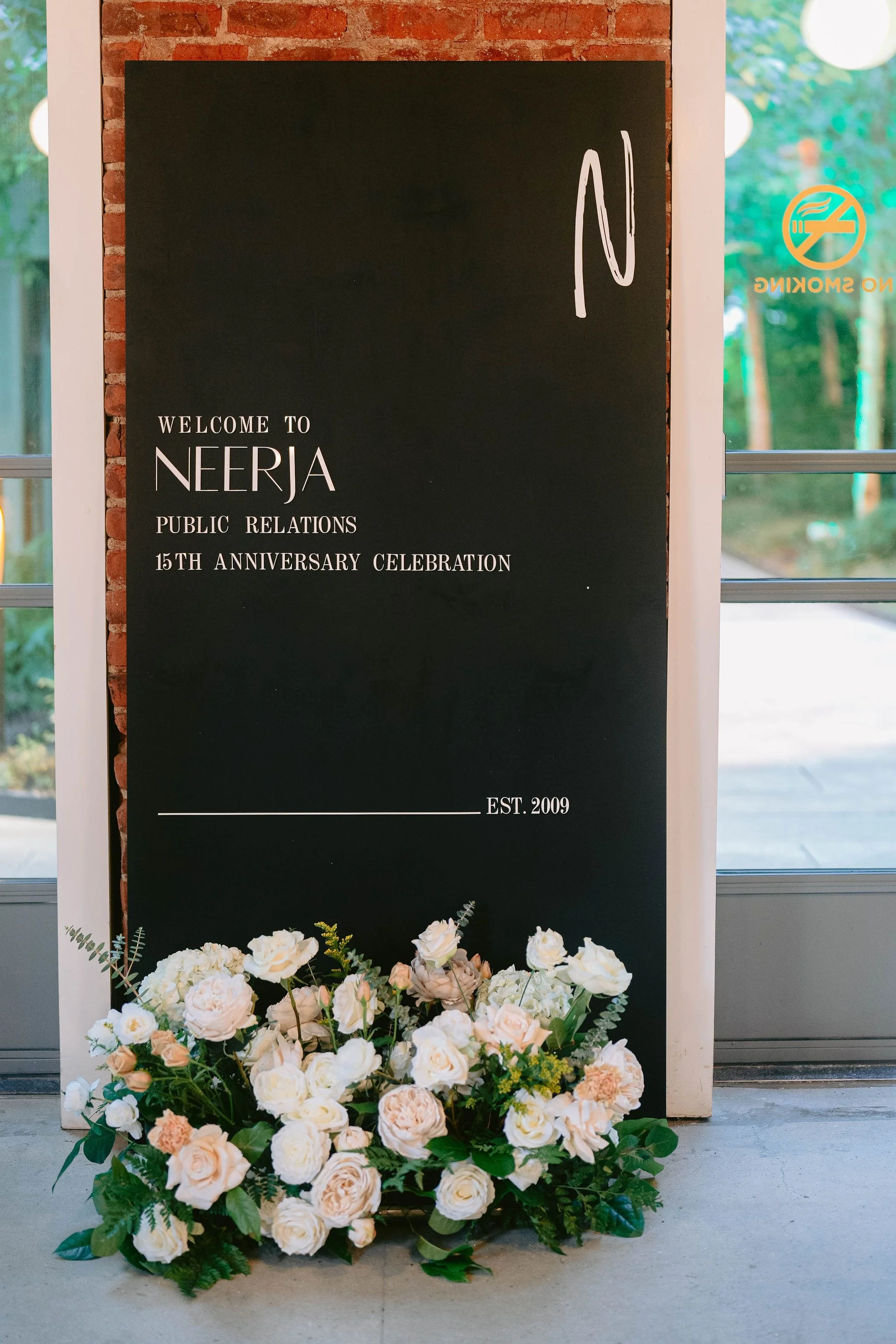 A floral arrangement of white and light pink roses with green leaves placed on the floor in front of a black poster board. The poster board displays the words 'Welcome to Neerja Public Relations 15th Anniversary Celebration' and the establishment yea