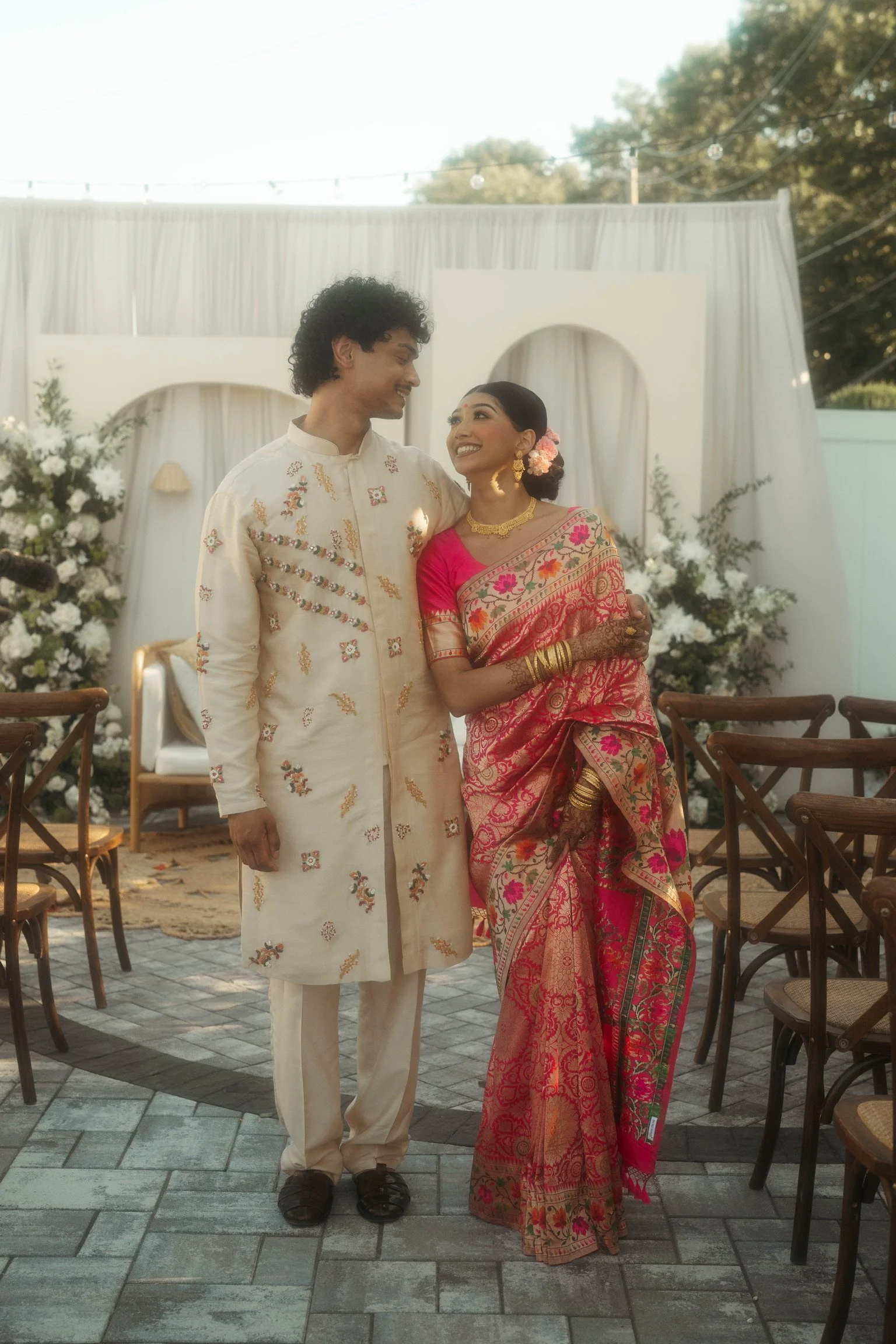 A man and woman dressed in traditional Indian attire standing close together, smiling at each other, at an outdoor wedding ceremony with white draped backdrop and floral arrangements.