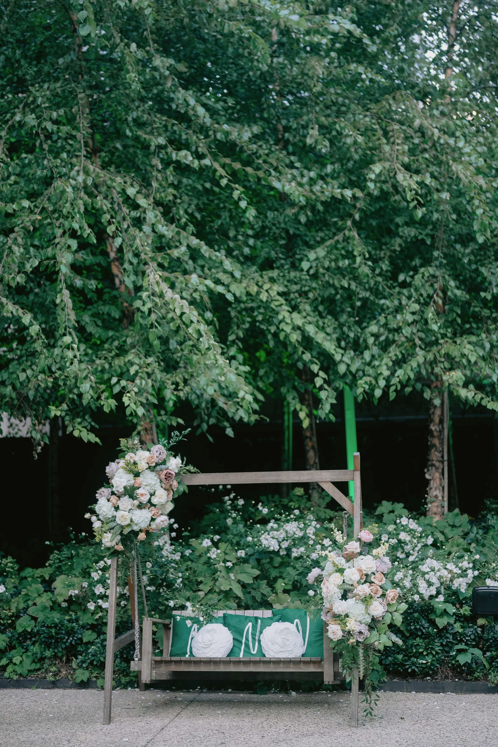 A wooden bench decorated with white and light pink flowers, greenery, and a green sign with white text; in front of lush green trees.