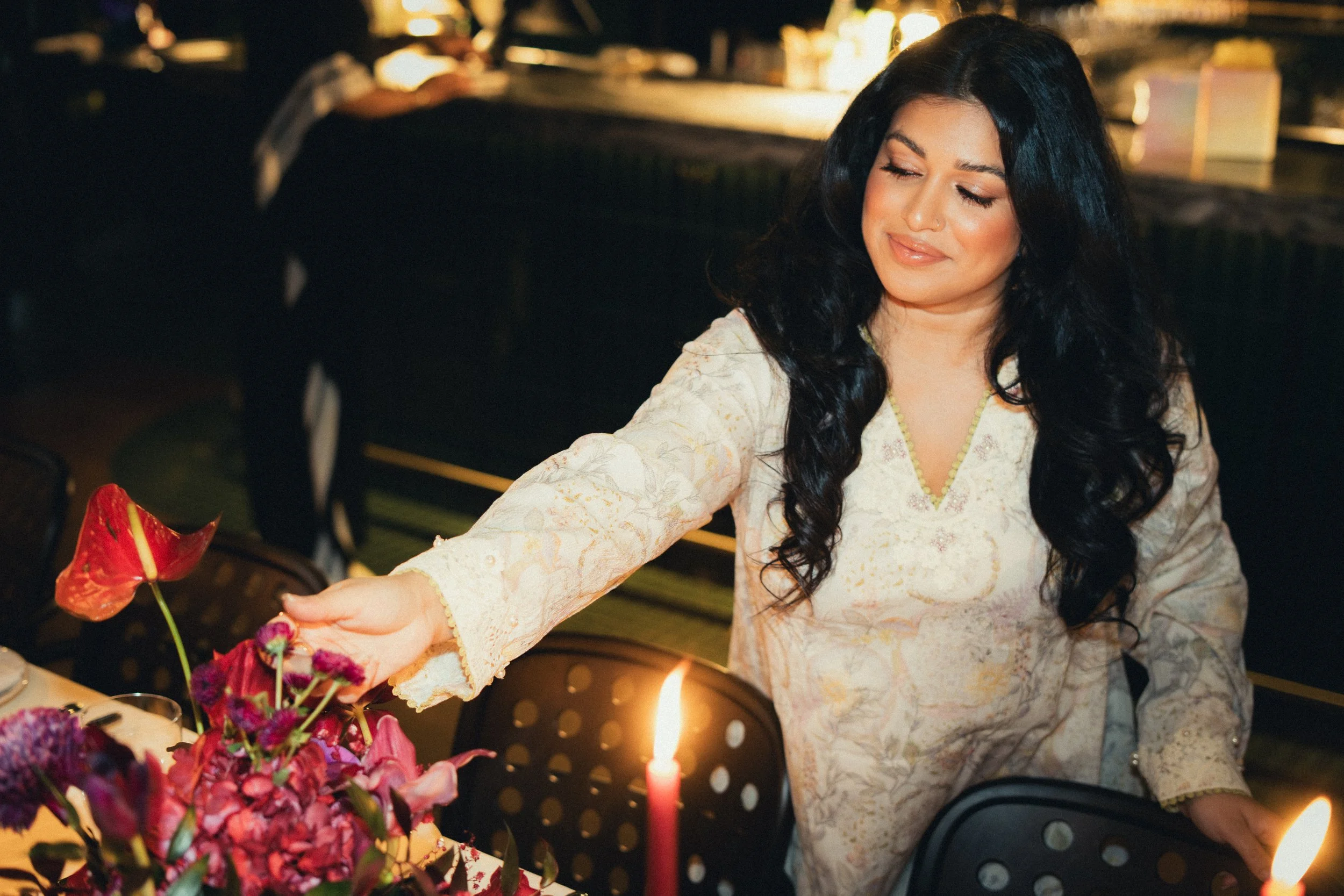 A woman wearing a light-colored traditional dress is placing a flower near a lit candle on a table with flowers, possibly during a celebration or ritual, in a dimly lit setting.