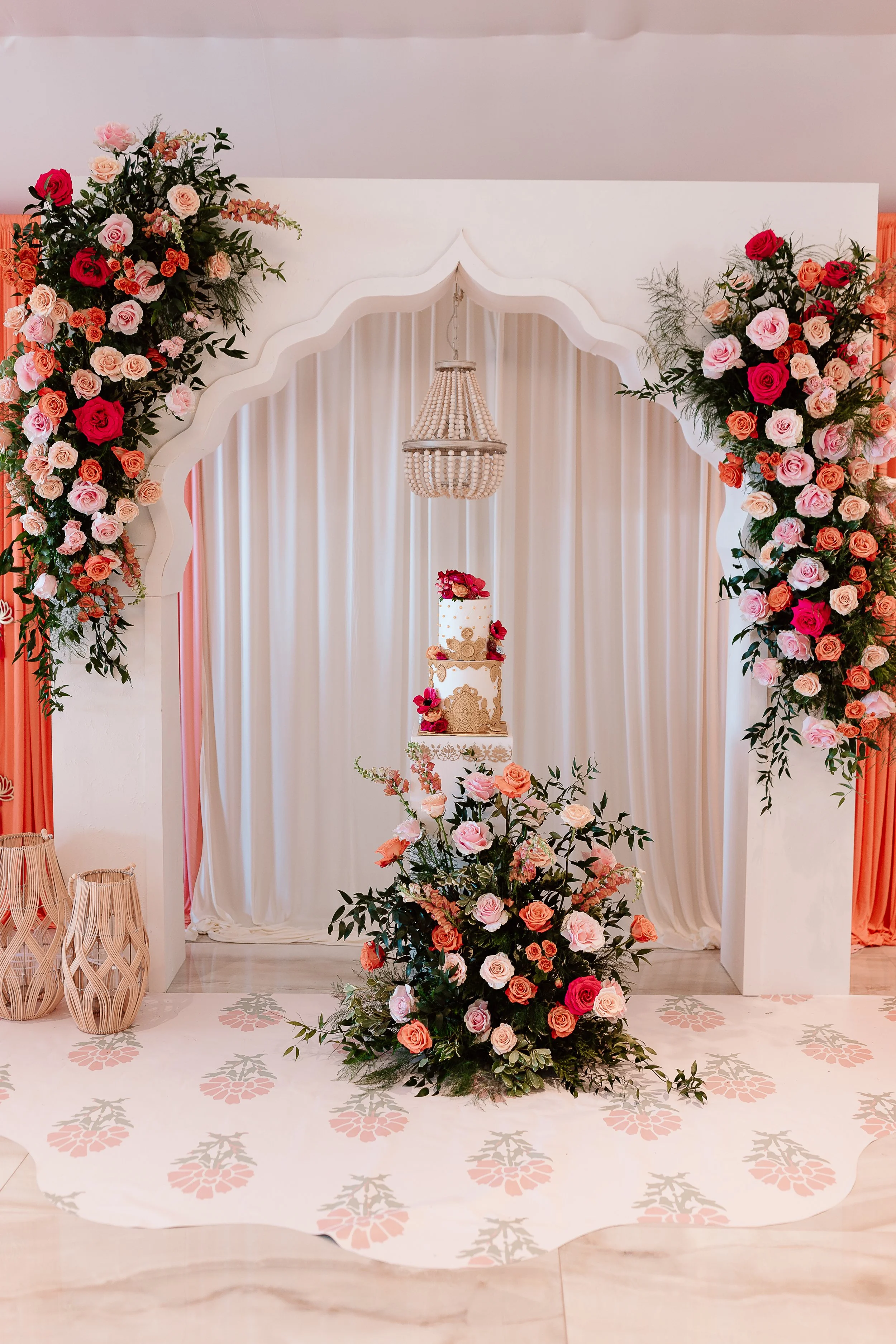 A decorated wedding or event backdrop featuring a floral arch with pink, peach, and red roses, a white ornate cake stand with a cake and flowers, suspended chandelier, and floral arrangements on the floor with matching colors, all against a light cur