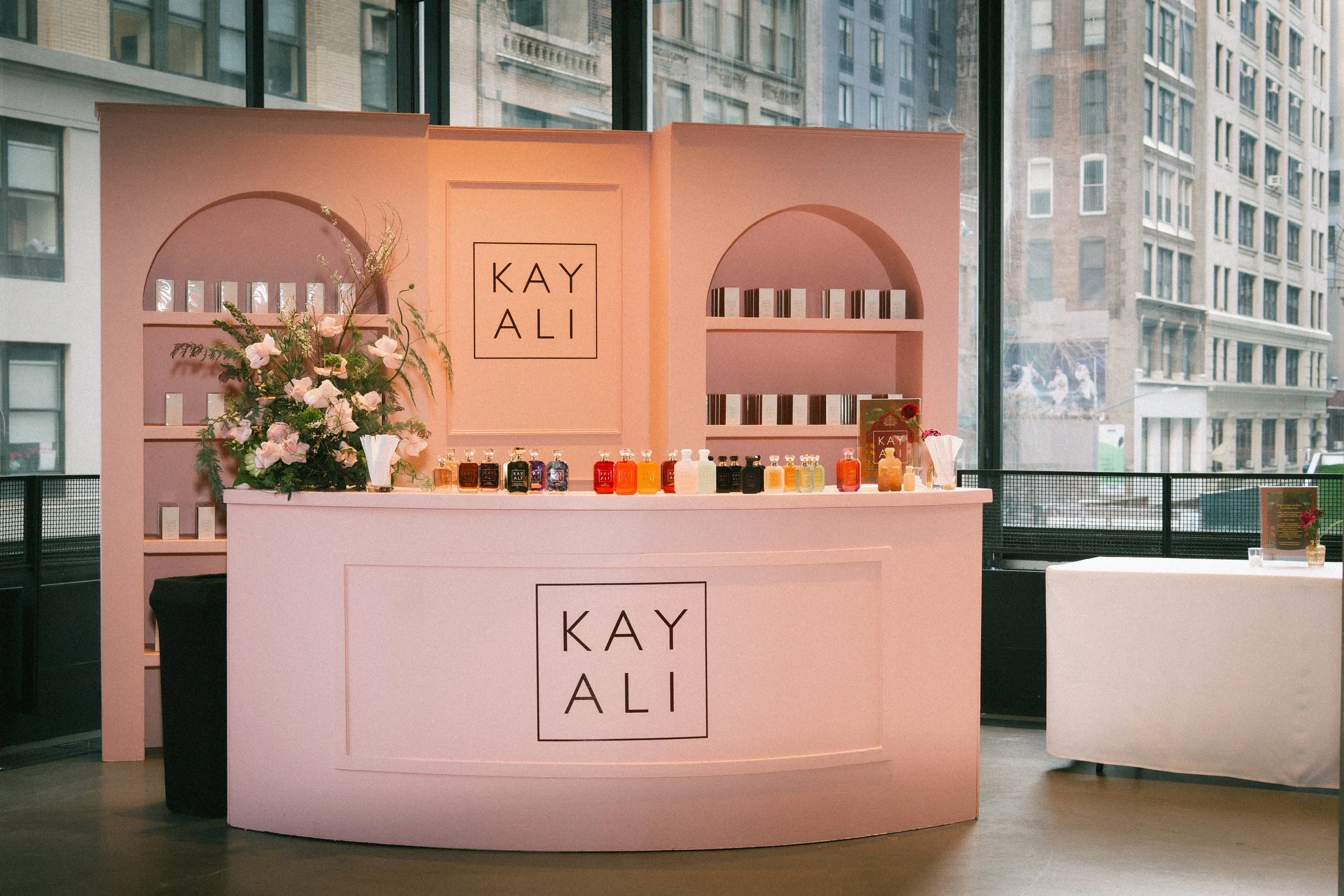 Pink display booth with the name 'KAY ALI' on it, decorated with flowers and bottles of various colored liquids, located inside a building with large windows and city buildings outside.