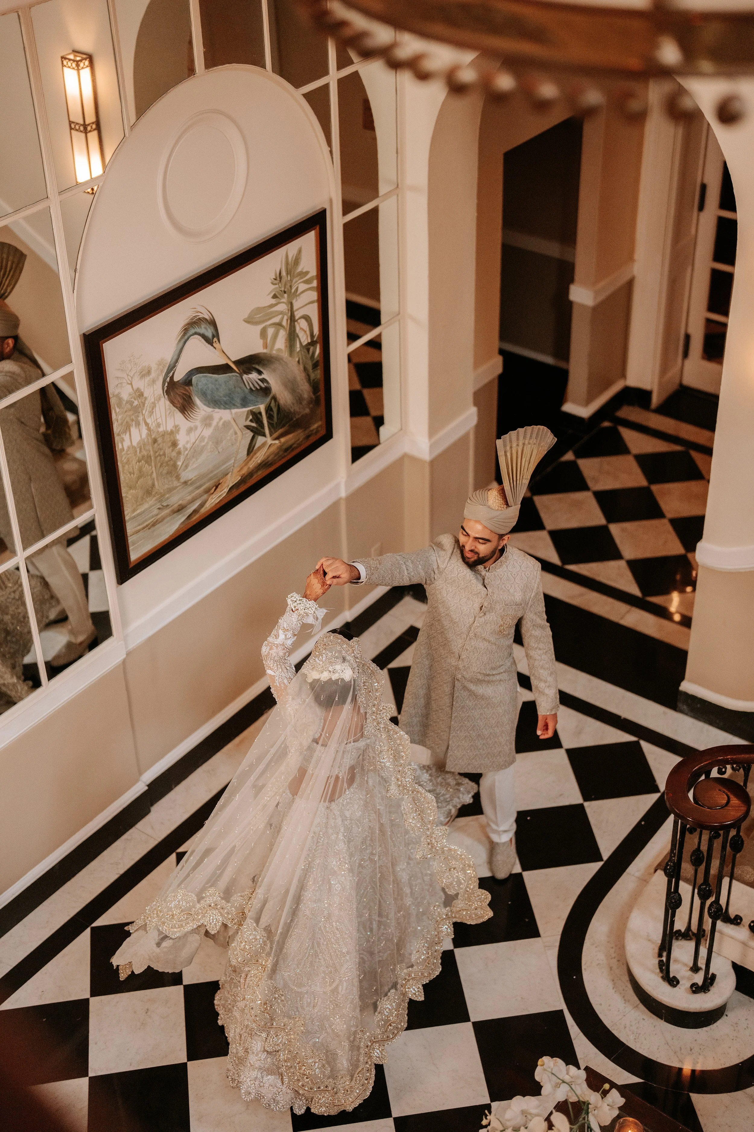 A bride and groom dancing at a wedding reception in a luxurious interior with black and white checkered floor, ornate railing, and framed artwork of a heron on the wall.