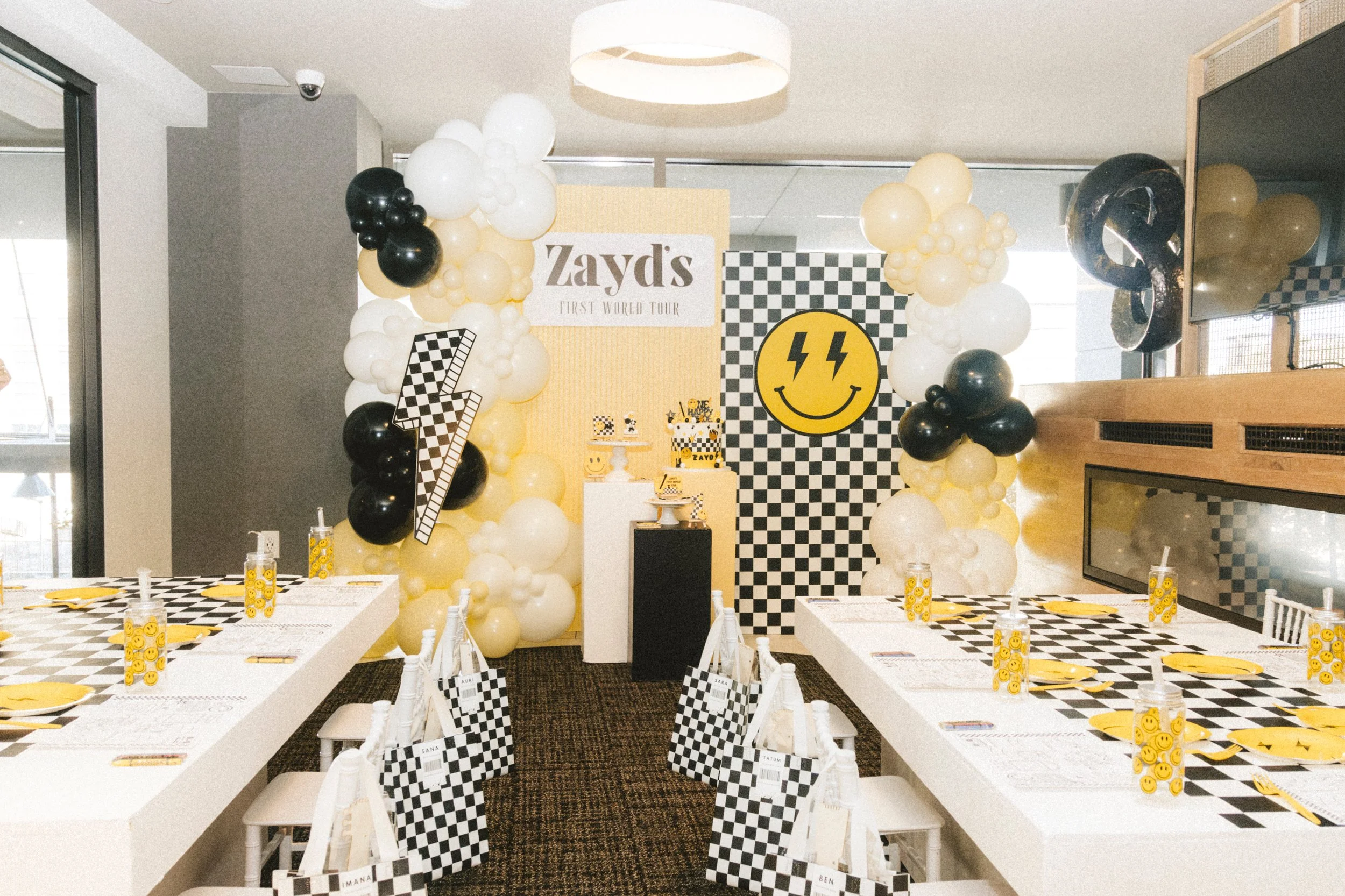 Party room decorated with black, white, and yellow balloons in an arch, with a backdrop that reads "Zayd's First World Tour" and features a smiley face with lightning bolts for eyes. Tables are set with yellow plates, smiley face themed cups, and che