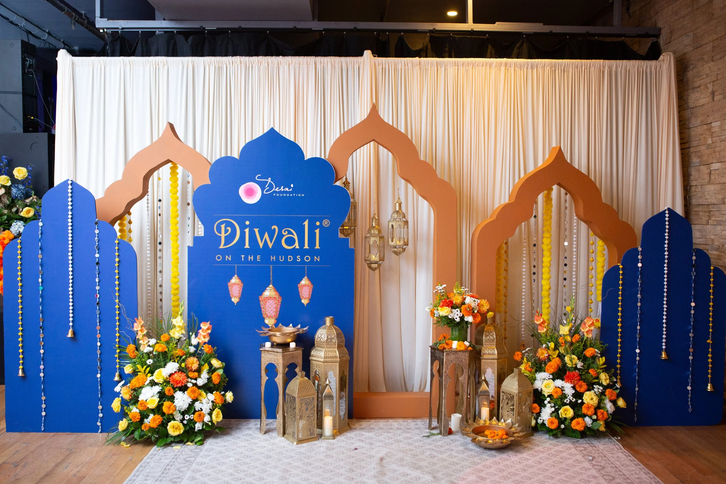 Decorative setup for Diwali celebration with colorful floral arrangements, lanterns, and ornate arches, featuring a blue sign with the text 'Diwali on the Hudson' in front of a cream-colored curtain backdrop.