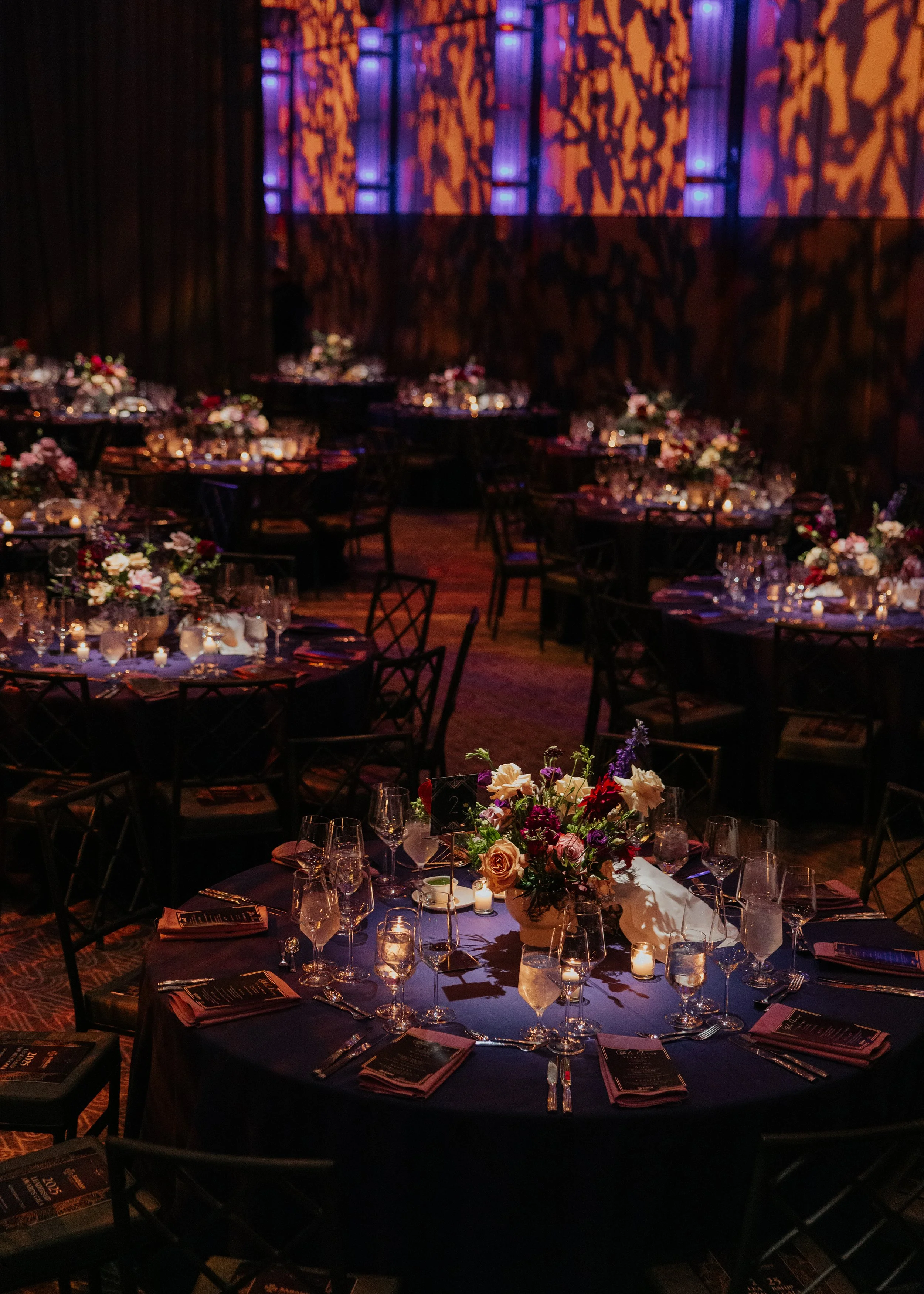 Elegant event space with decorated tables featuring floral centerpieces, candles, glassware, and cutlery, illuminated with soft lighting and backlit patterned wall panels.