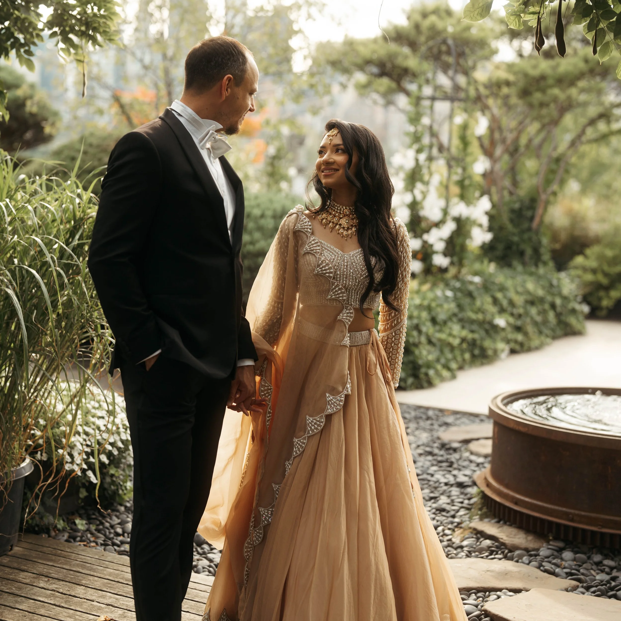 A couple dressed in traditional wedding attire standing outdoors among trees, holding hands, and smiling at each other.