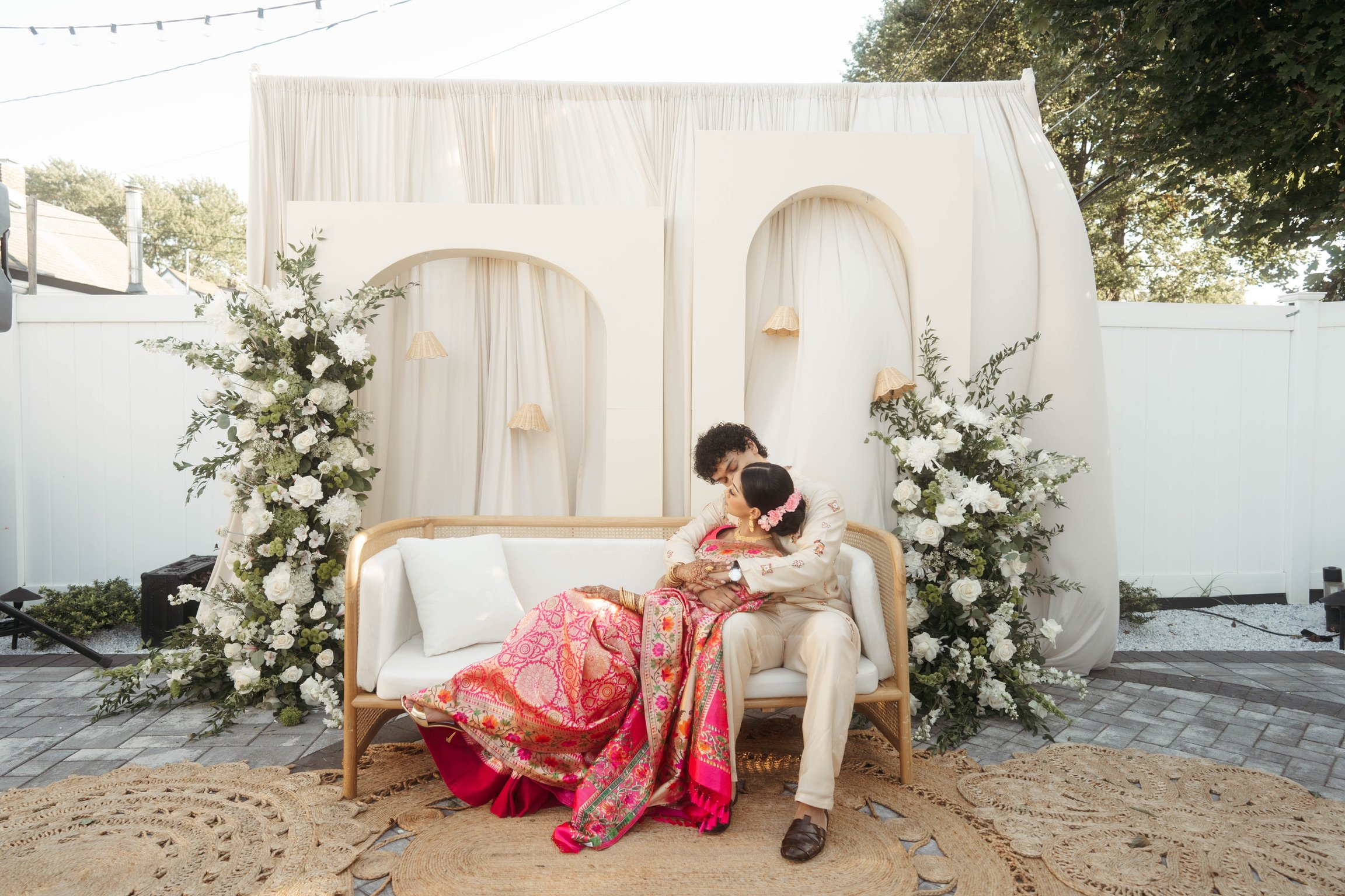 A couple dressed in traditional Indian attire sitting on a cream-colored sofa decorated with large floral arrangements and white curtains, during an outdoor celebration.