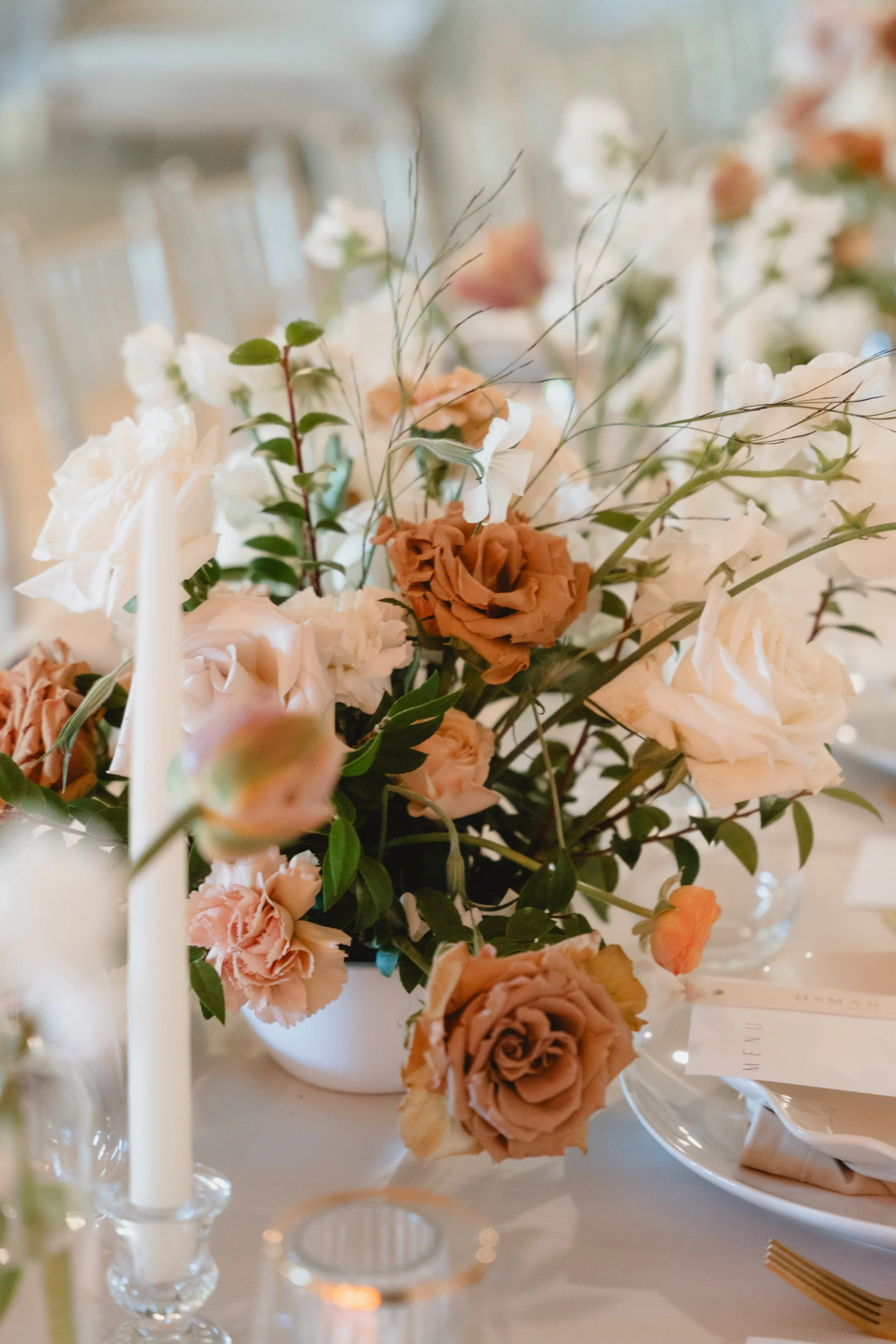 A floral arrangement with roses and greenery in a white vase on a table with candles and place settings.