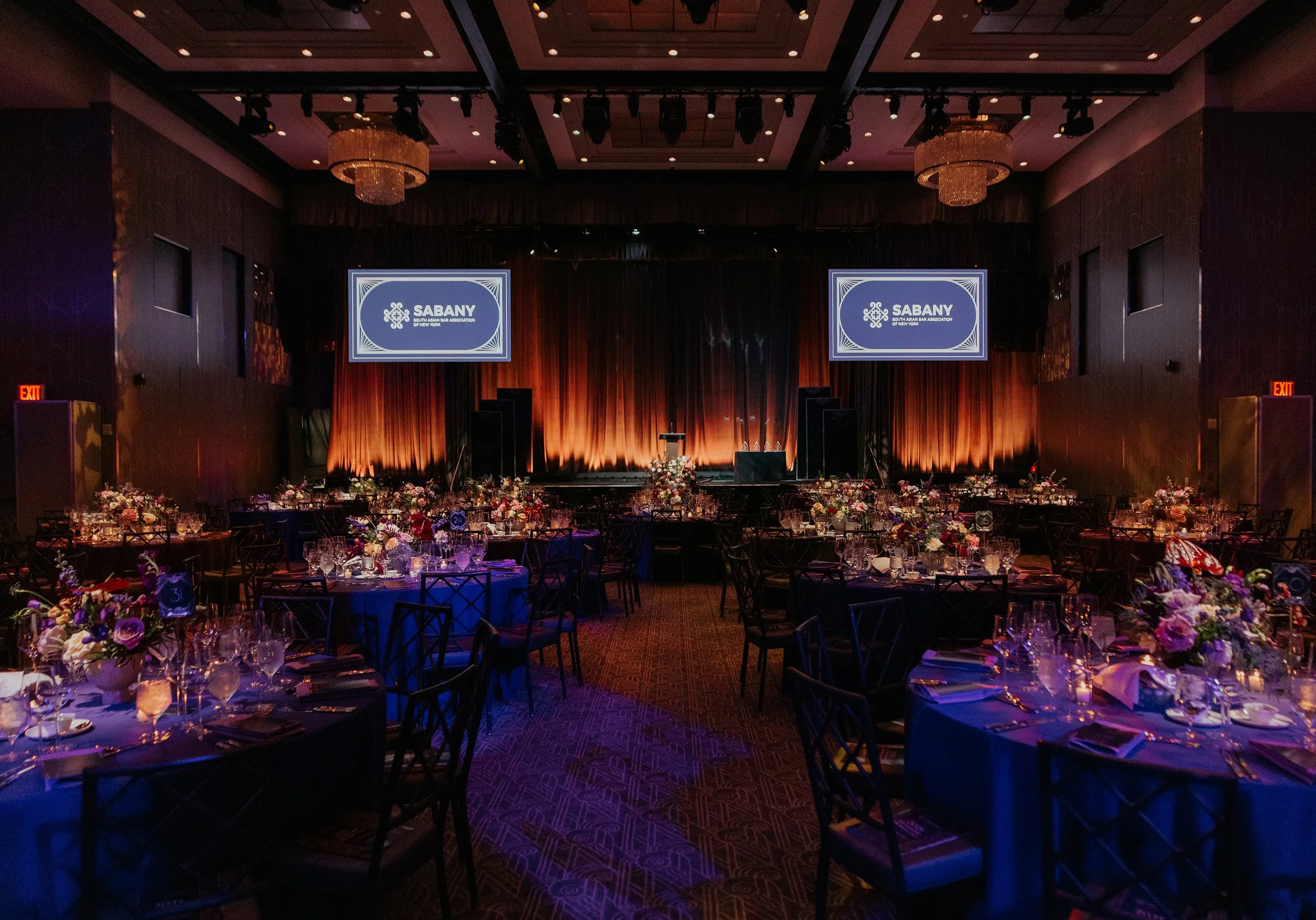 A large banquet hall with decorated tables, floral centerpieces, and a stage with two large screens displaying 'SABANY South Asian Bar Association of New York.' The room is dimly lit with warm lighting.