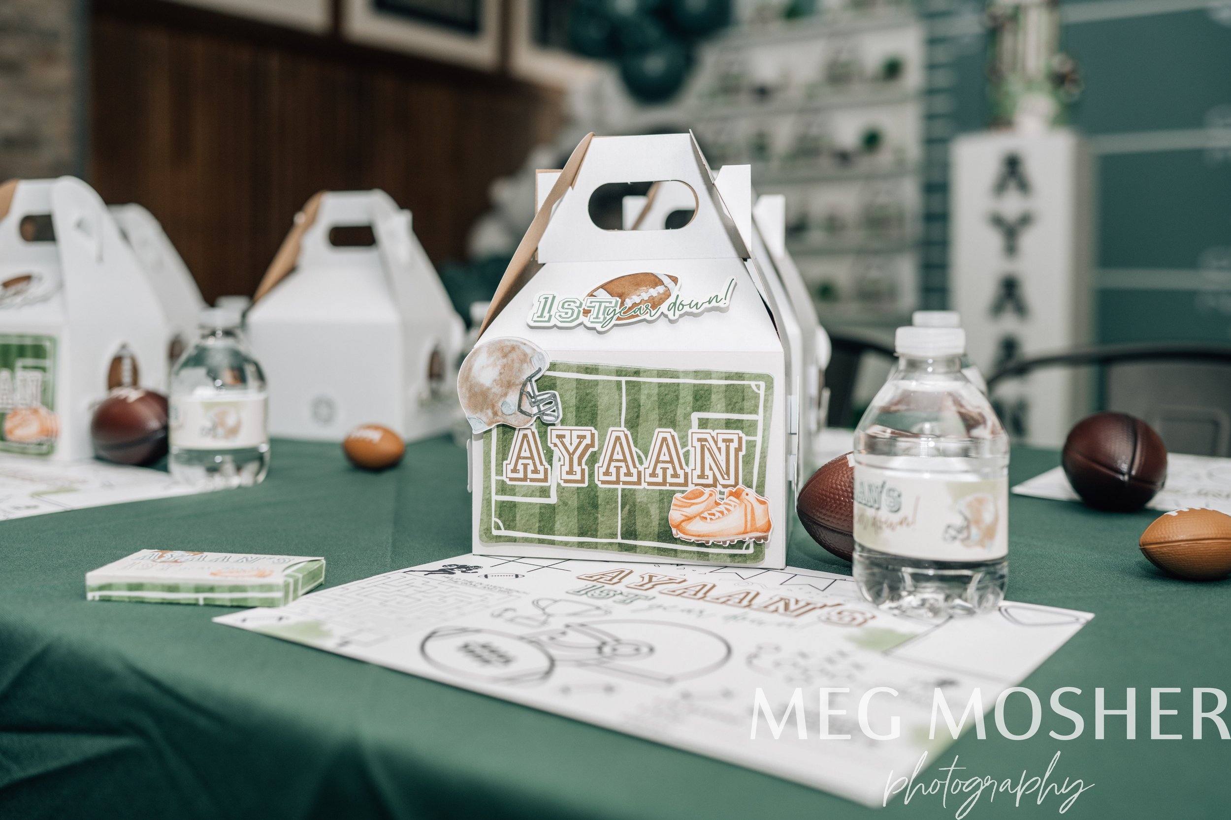 A green table with football-themed party favors including a decorated cake box with football graphics and the name 'Ayaan', various football-shaped candies, and water bottles. In the background, blurred shelves with additional party supplies.
