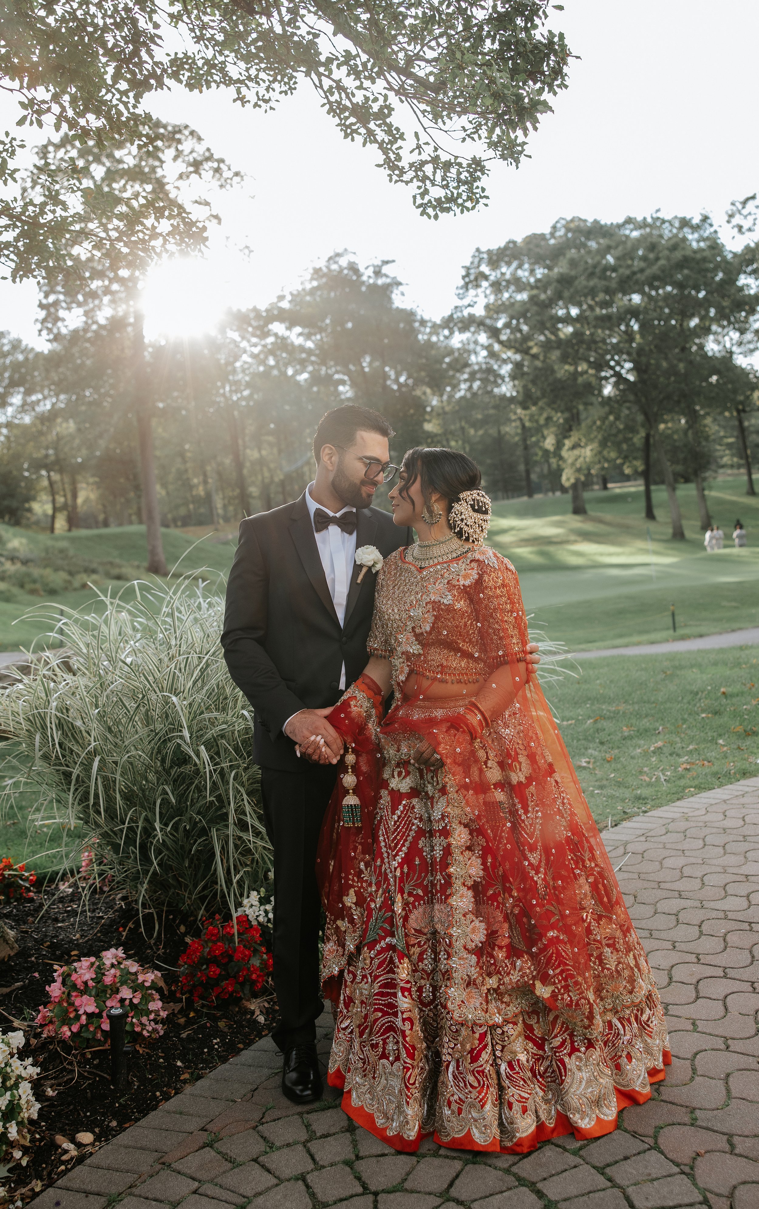 A couple dressed in traditional and formal wedding attire, standing outdoors on a sunny day, surrounded by greenery and flowers, holding hands and gazing at each other.