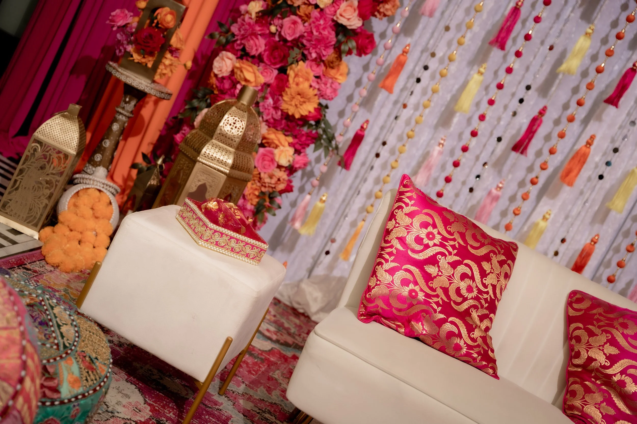 A decorative setting with pink, orange, and red flowers, lanterns, and colorful hanging tassels on a wall. Two white chairs with ornate pink and gold pillows are placed in front of the display. The scene appears to be part of a festive or celebratory