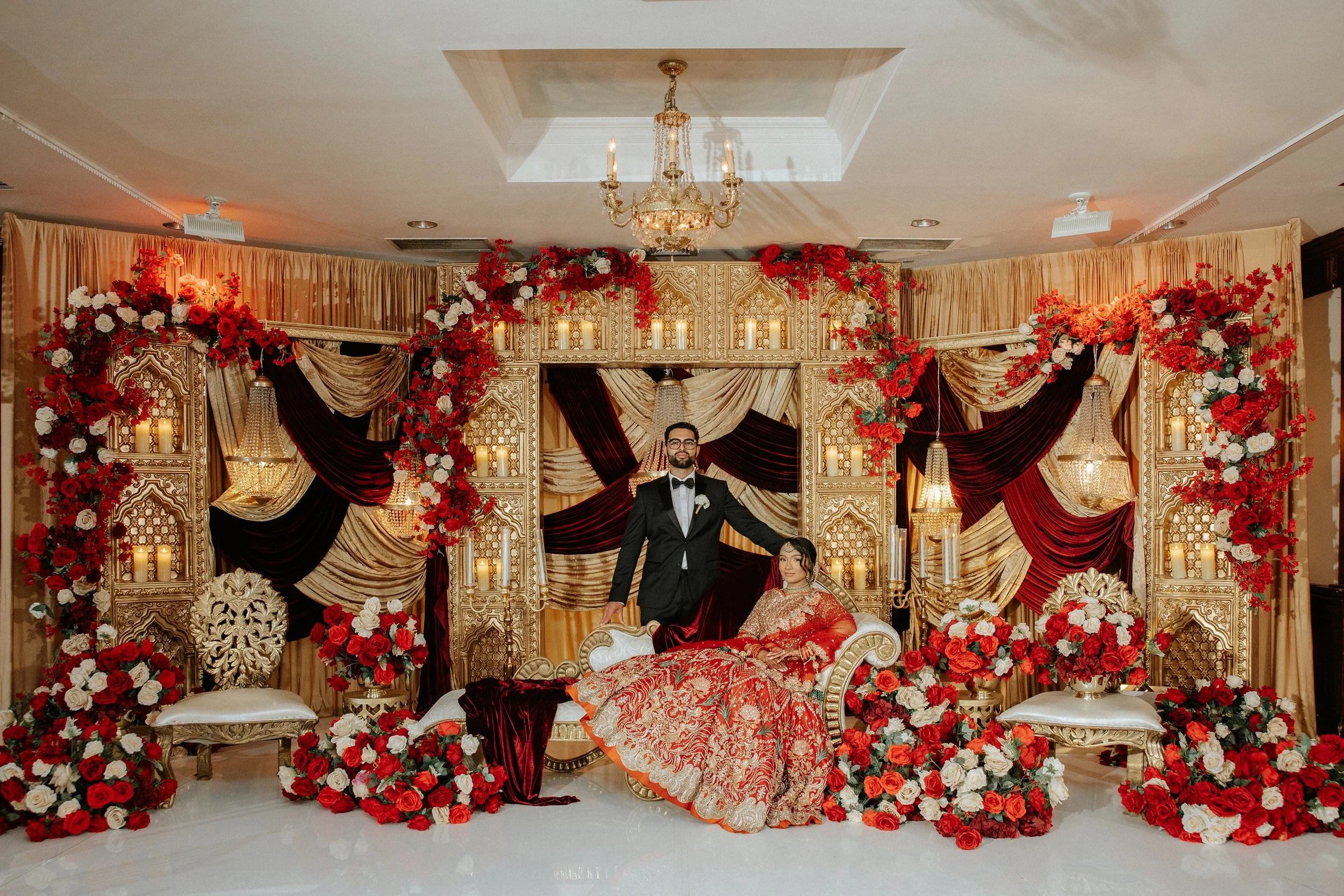 A decorated stage for a wedding with red and white flowers, gold drapes, and a chandelier. A couple dressed in traditional wedding attire is posing, with the groom standing and the bride sitting on a luxurious sofa.