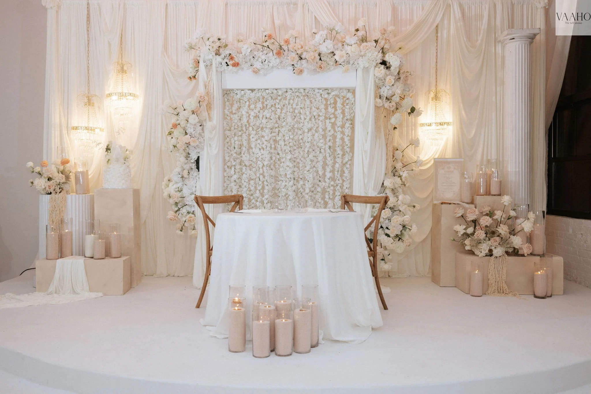 Wedding backdrop with white drapery, floral arrangements, and candles, set with a small table and two chairs in a neutral color scheme.