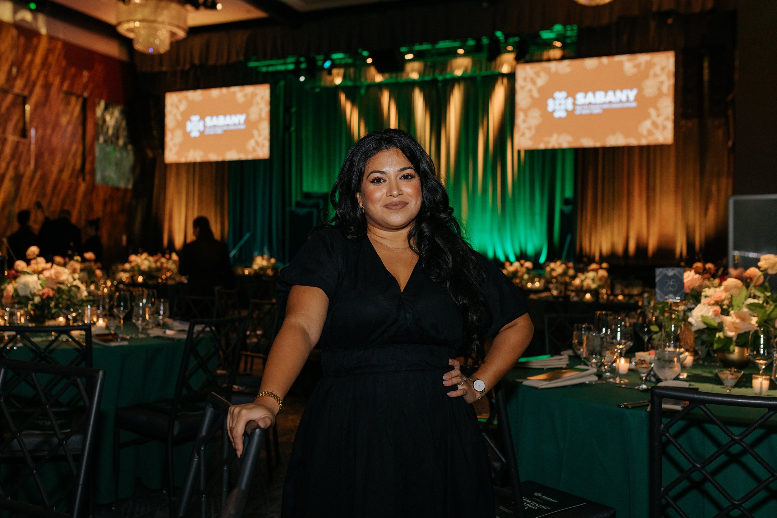 A woman with long dark hair wearing a black dress and watch, standing in a decorated event hall with tables, flowers, and candles.