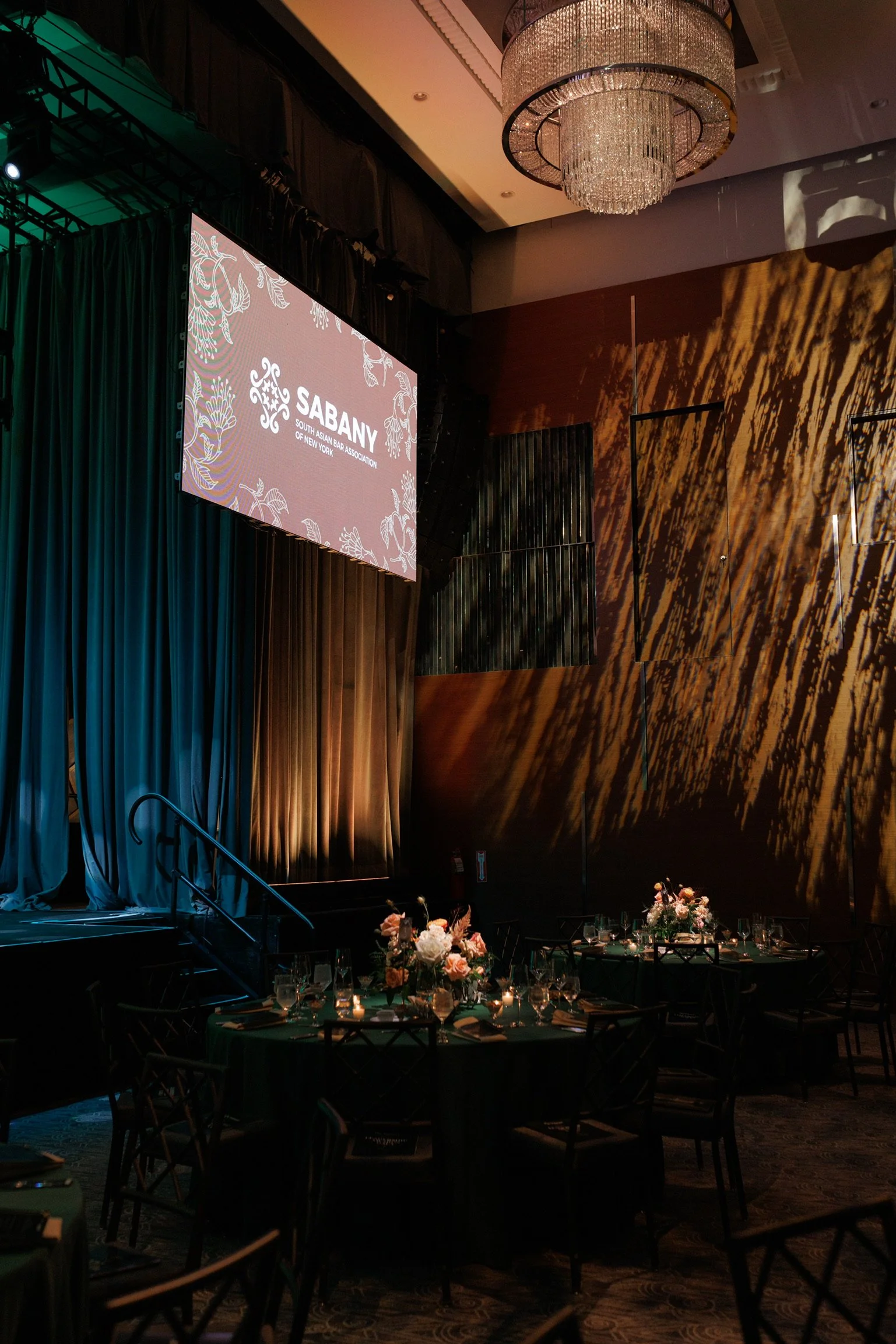 An elegant event space with a large chandelier, a stage with curtains, a projection screen displaying the Saban logo for the South Asian Bar Association of New York, and decor with tables set for a formal dinner with floral centerpieces and candles.