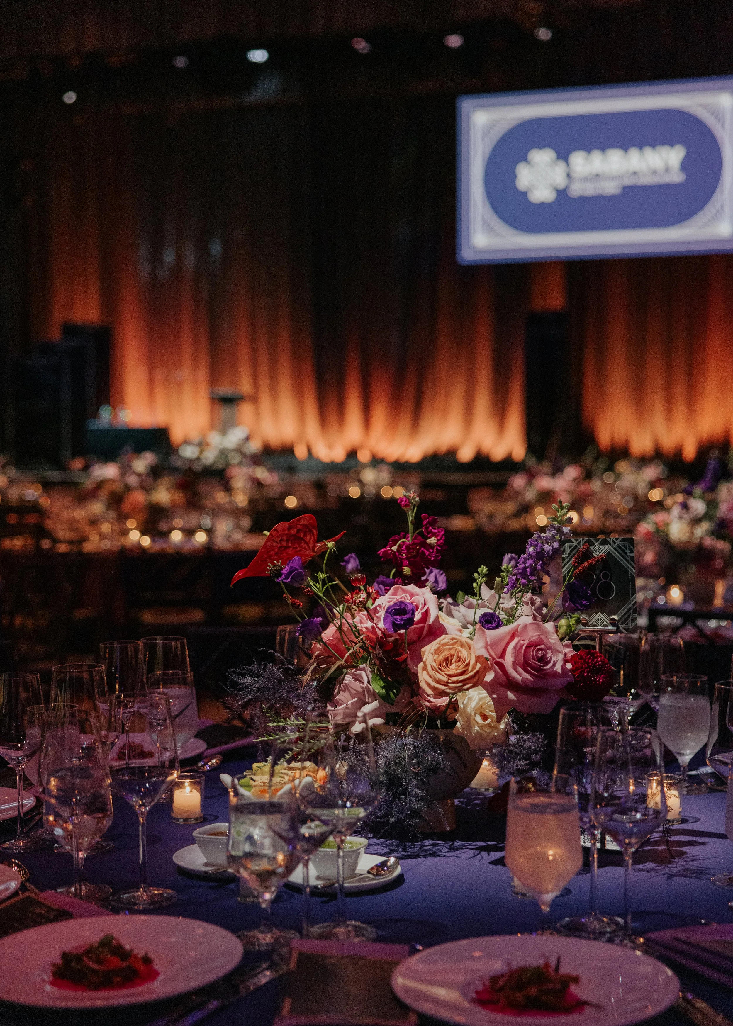 An elegant banquet table with a large floral centerpiece consisting of pink, purple, red, and peach flowers, surrounded by glasses, candles, and plates, with a stage and a large screen displaying the SABNY logo in the background.