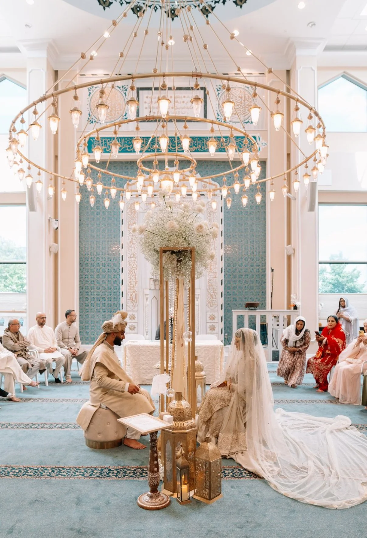A wedding ceremony inside a decorated hall with a high ceiling, featuring elegant chandeliers. The bride and groom are seated in traditional attire, facing each other, with guests seated on either side. The background has intricate decor and floral arrangements, including a large bouquet of white flowers.