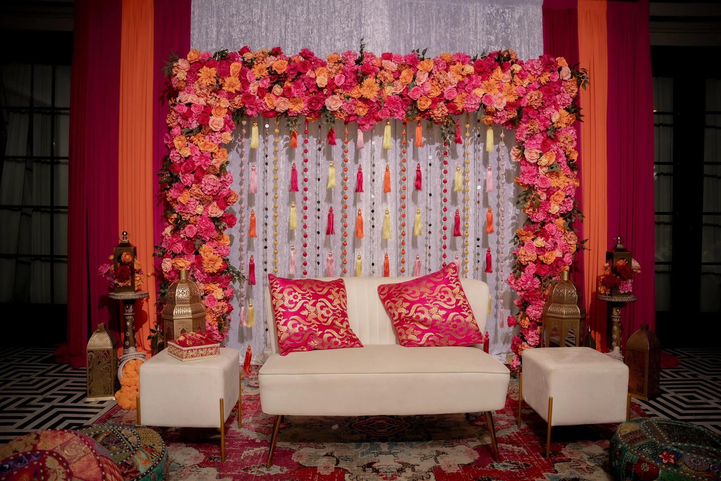 A decorative backdrop with pink, orange, and peach flowers, tassels, and beads. In front, there is a white sofa with two pink and gold patterned pillows, and two white ottomans. Two gold lanterns with pink flowers on top are on either side.