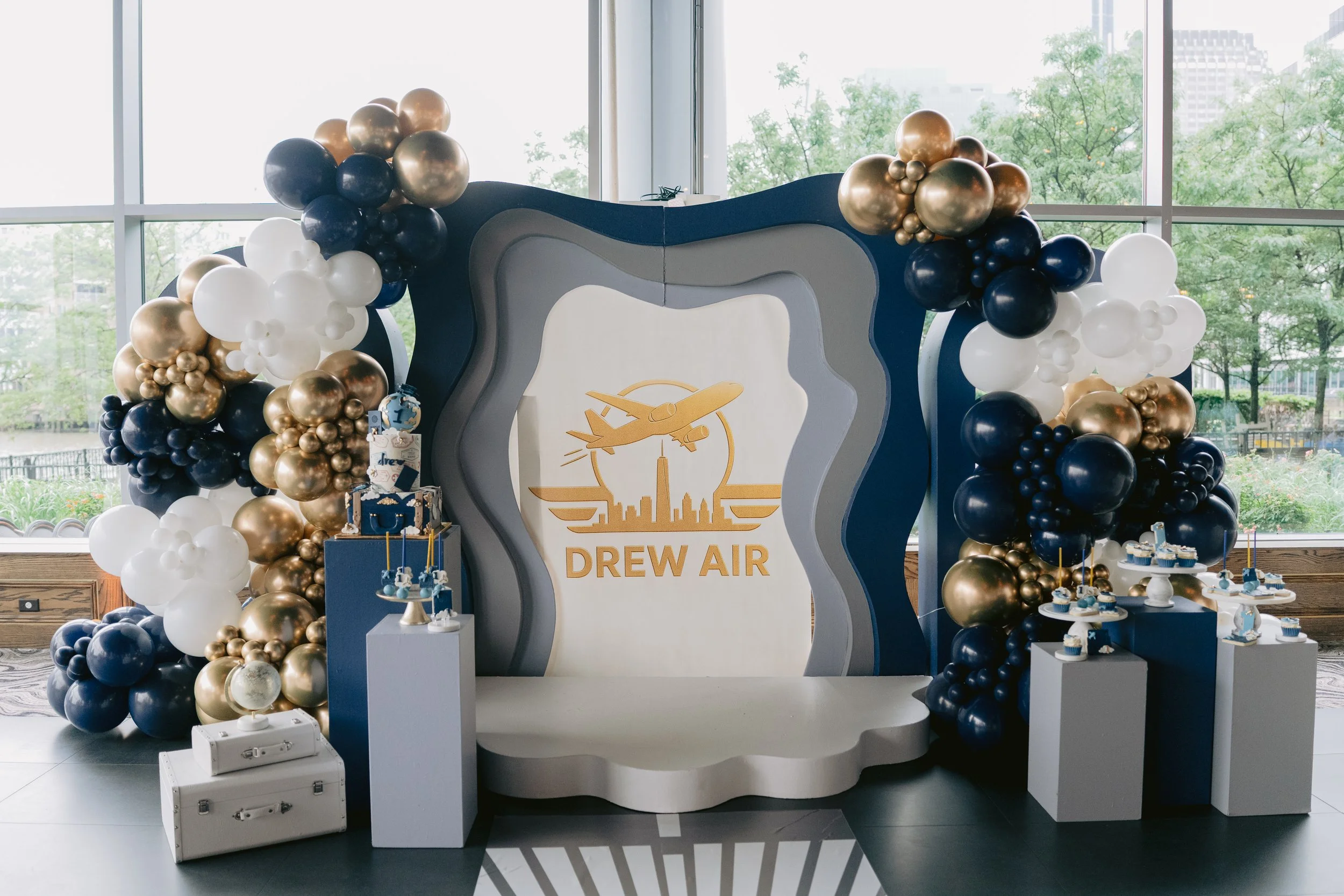 Decorative display for Dreww Air with a backdrop featuring a stylized airplane and cityscape graphic, surrounded by clusters of balloons in navy blue, gold, and white, with small dessert tables holding cupcakes and treats adjacent to white trunks and
