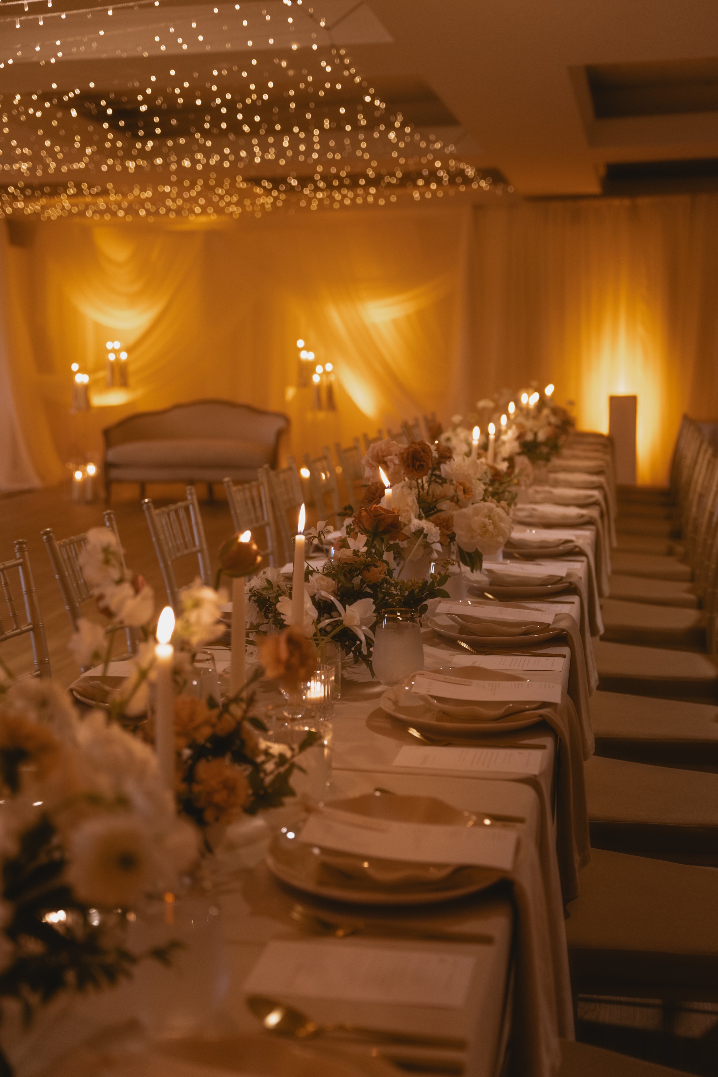 Elegant banquet hall with warm lighting, decorated with floral centerpieces, candles, and set dining tables.