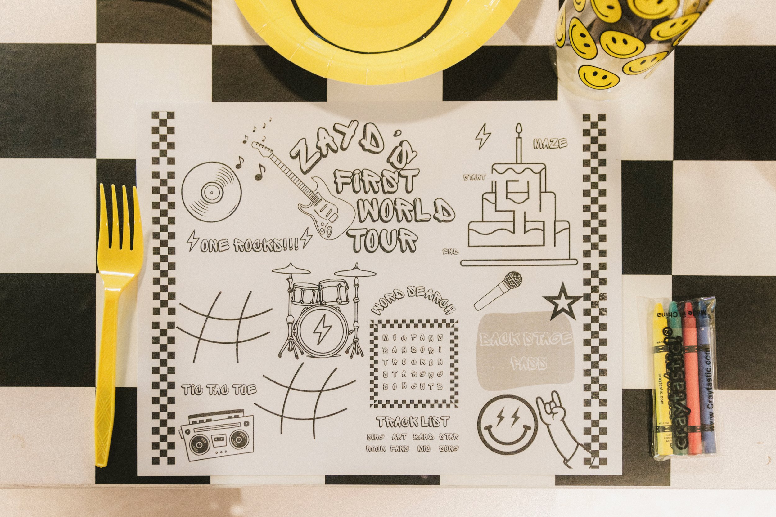 Coloring sheet with drawings and words related to a music tour, with a yellow plate, yellow smiley face cup, yellow fork, and a pack of colorful Crayola markers on a black-and-white checkered surface.