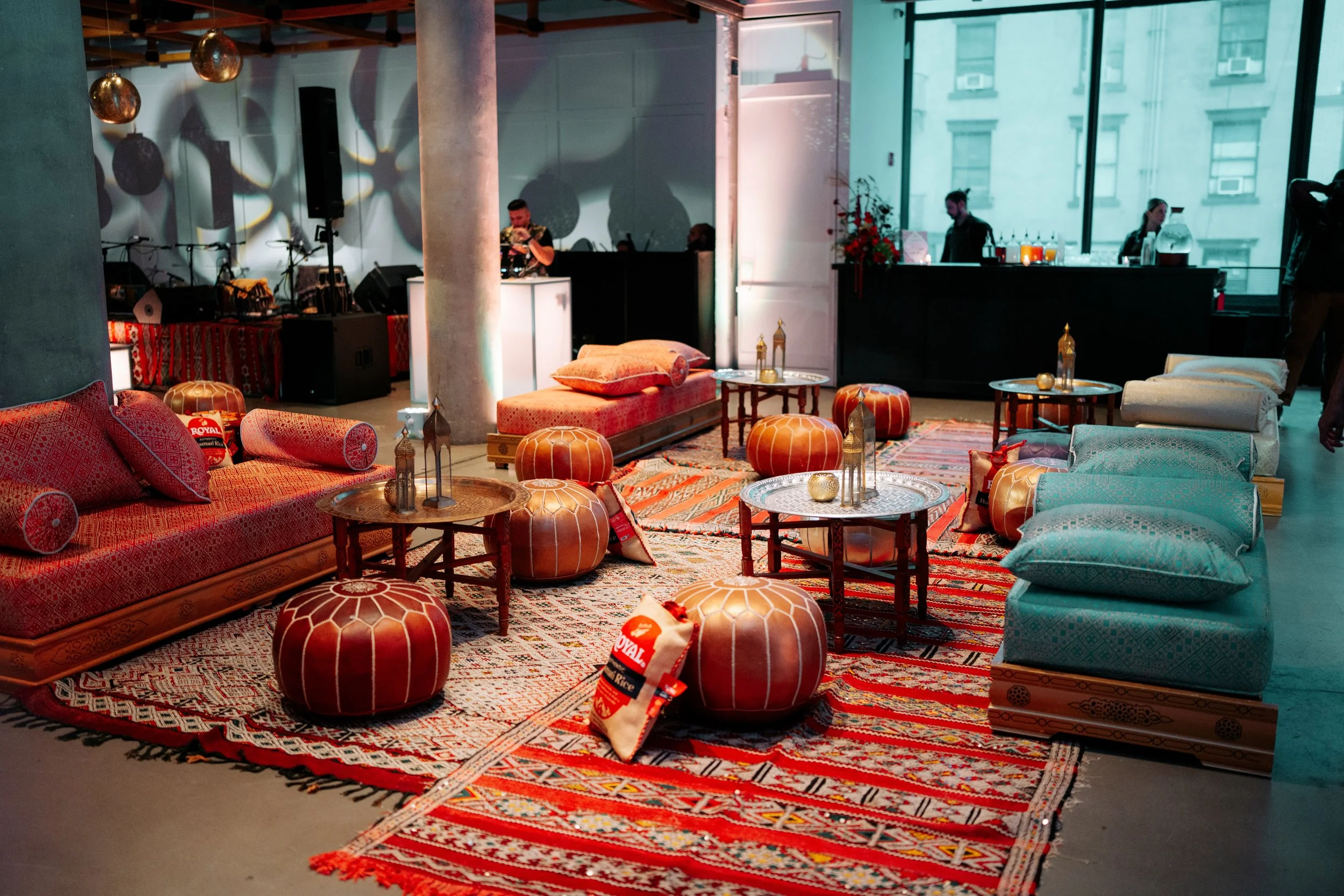Interior of a modern living space with colorful rugs, upholstered seating, and poufs, featuring a stage with musical instruments and a DJ setup, decorated with hanging lights and lanterns, in front of large windows overlooking an urban building.