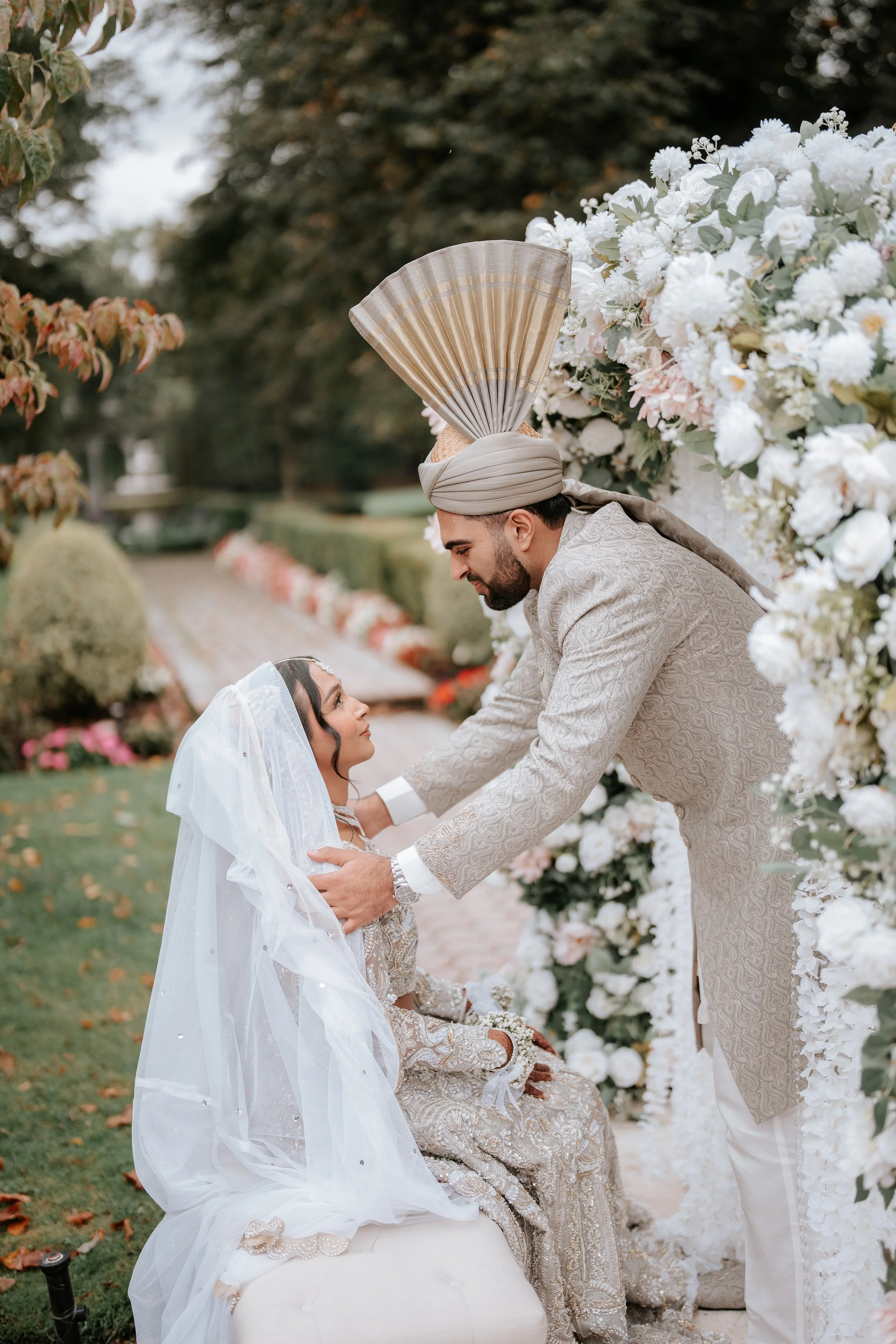 A groom and bride at a wedding with a floral arch and garden background. The groom, wearing a beige embroidered sherwani with a turban, is kneeling and holding the bride's hands. The bride, dressed in a white wedding dress with a veil, is sitting and