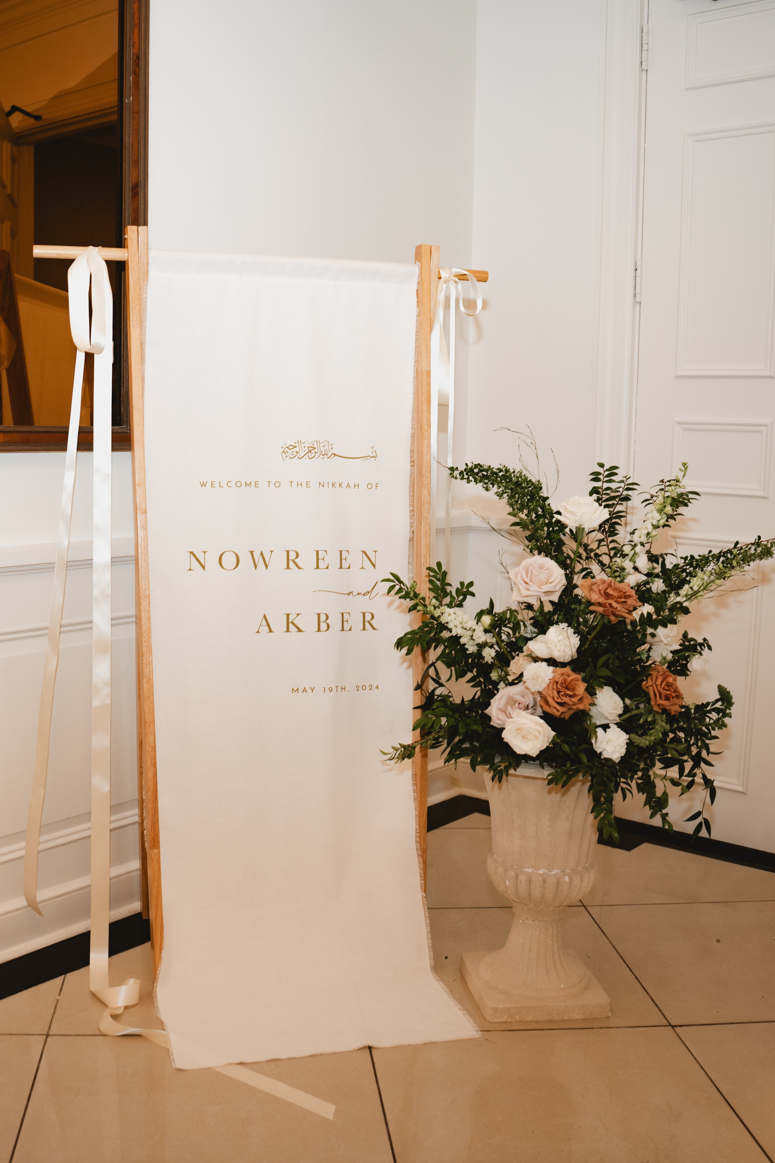 A white wedding welcome sign with gold lettering and decorative script, placed on a wooden easel, next to a large floral arrangement in a beige urn, indoors with white paneled walls and tiled floor.